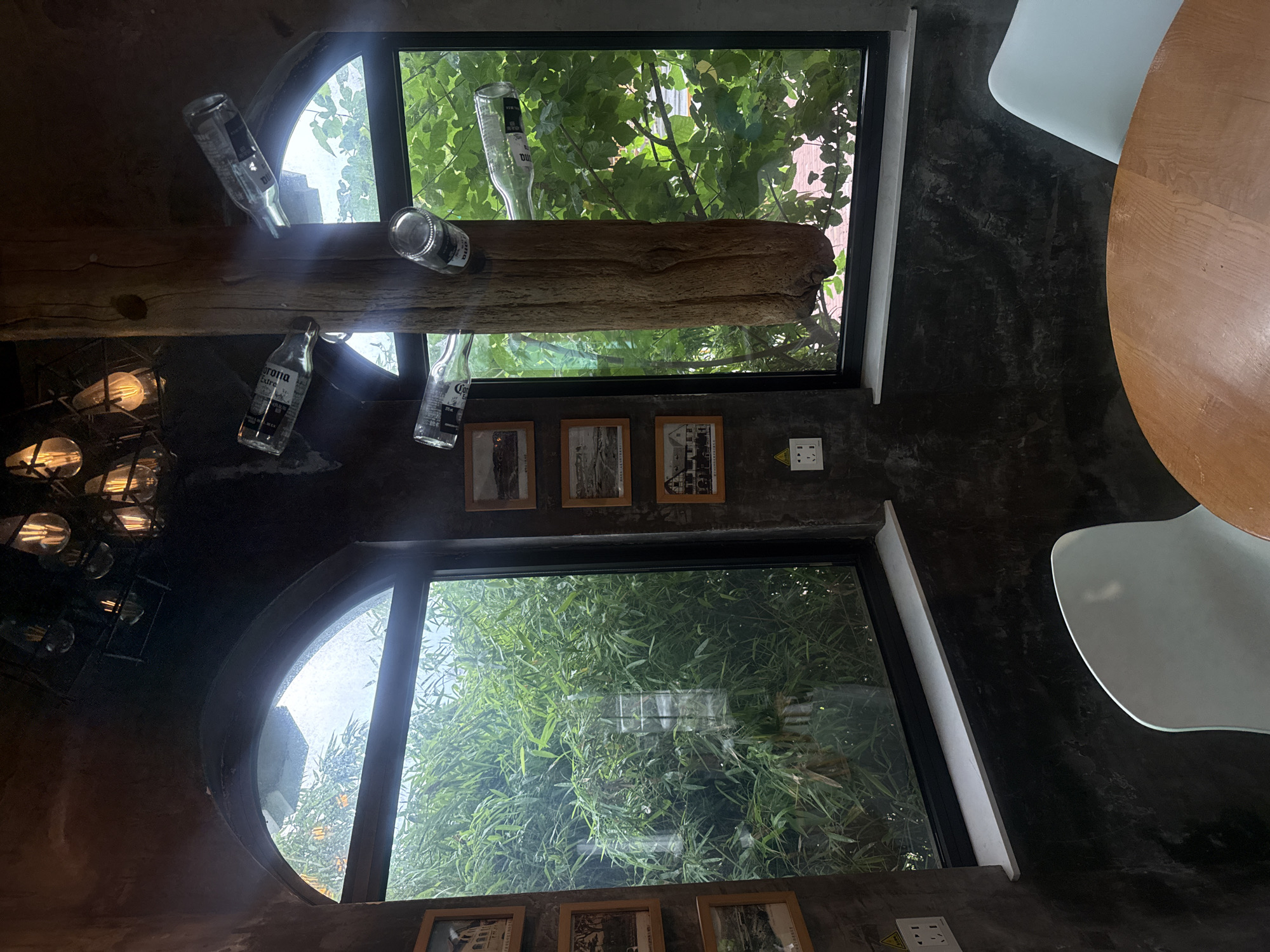 The image depicts an indoor scene featuring two large windows with arched tops, looking out onto lush greenery. Several glass bottles, seemingly repurposed as decorative elements, are suspended or affixed to a thick, rough-hewn wooden beam that stands vertically between the two windows. These bottles appear to be beer bottles, with labels visible on some. Above the windows, a cluster of industrial-style light fixtures with exposed bulbs hangs from the ceiling. On the wall to the left of the left window, a series of framed photographs are displayed. Electrical outlets are visible near the bottom of the left window and on the wall to the right of the right window. In the foreground, a portion of a round wooden table and a white chair are visible. The overall ambiance suggests a rustic or industrial-chic interior design, possibly a cafe or restaurant. The provided image does not contain any information or visual cues that would allow for the determination of its specific location within Qinhuangdao, China, or any other city.