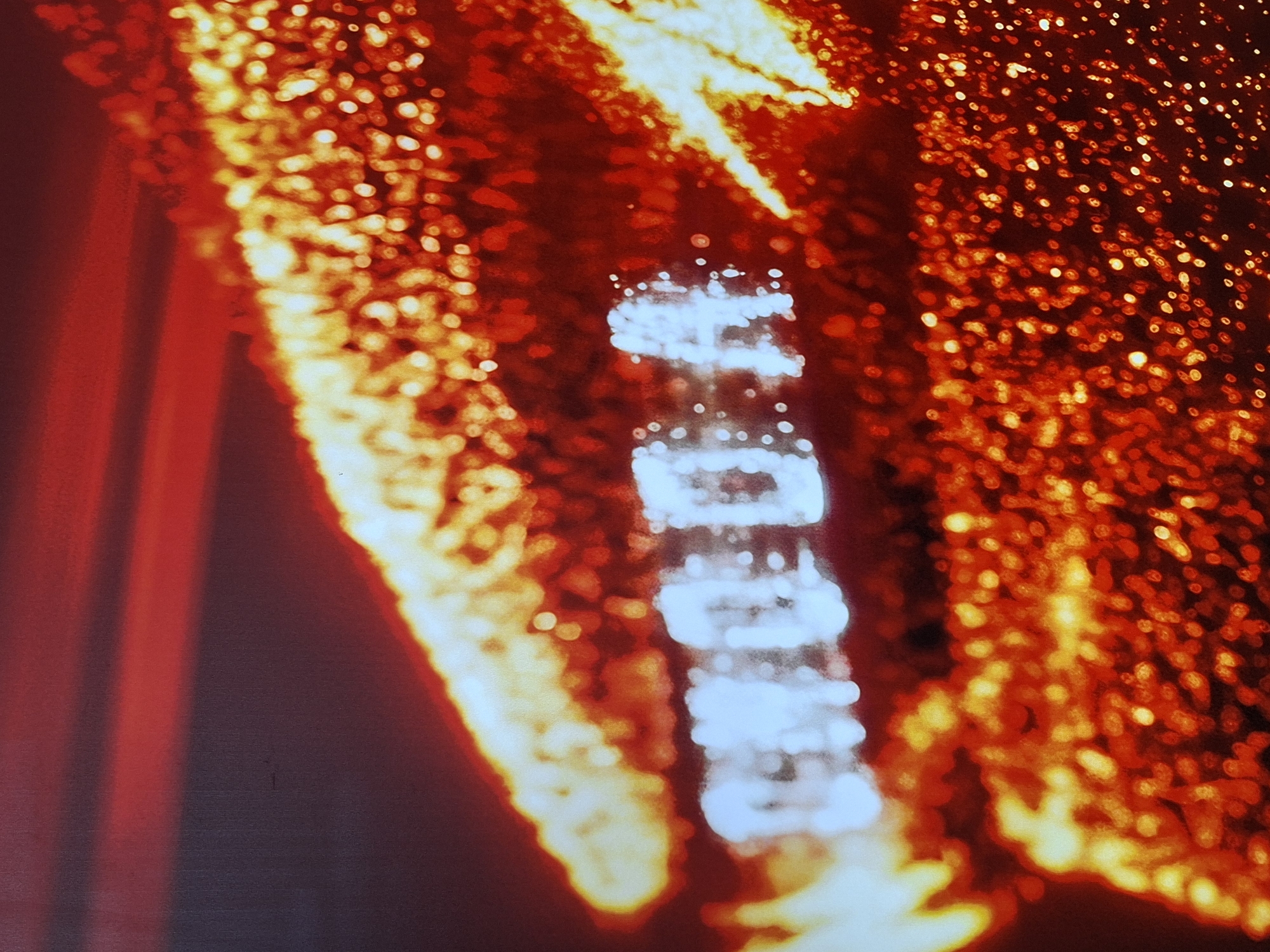 The image is a close-up shot of a sign that reads "CHODA" in white, illuminated lettering against a blurred background of red and orange lights. The lettering appears slightly distorted due to the shallow depth of field and the speckled texture of the sign. There is no visible information in the image that allows for identification of its location in Izhevsk, Russia. The overall scene is abstract and focuses on the glowing text and the bokeh effect of the lights.