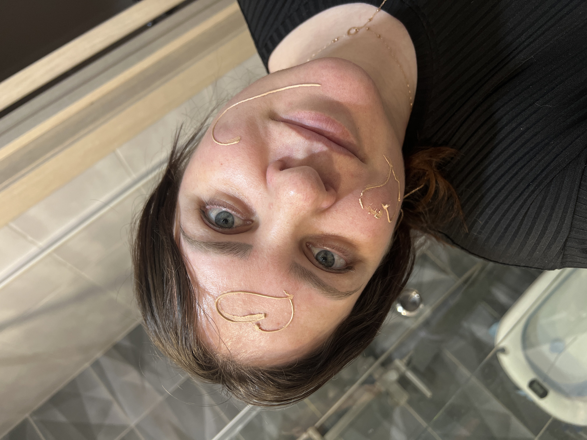 A fair-skinned woman, likely in her 30s or 40s, is captured in a close-up, chest-up shot. She appears to be lying on her back, looking directly at the camera with light blue-green eyes and a neutral, slightly inquisitive expression. Her dark hair frames her face. She wears a black, ribbed garment and a delicate gold-colored necklace with small, linked elements around her neck.
The notable detail is the presence of several thin, gold-colored, malleable pieces carefully arranged on her face. A single curved line rests on her forehead. On her right cheek (viewer's left), a long, curved line extends across her temple, and below it, a heart shape is formed. On her left cheek (viewer's right), two intertwined, wavy lines are visible. These pieces suggest an artistic application, temporary facial decor, or a beauty treatment.
The background indicates an indoor setting. In the upper left, wooden-toned panels or beams are visible. The lower background is reflective, possibly a glass surface or polished tiles, displaying patterns of light and dark grey and beige squares. An indistinct, light-colored object, potentially a basin or equipment, is partially visible in the bottom right. There is no visible text. The overall scene suggests a posed shot within a clean, modern interior, located in Noviy, Russia.