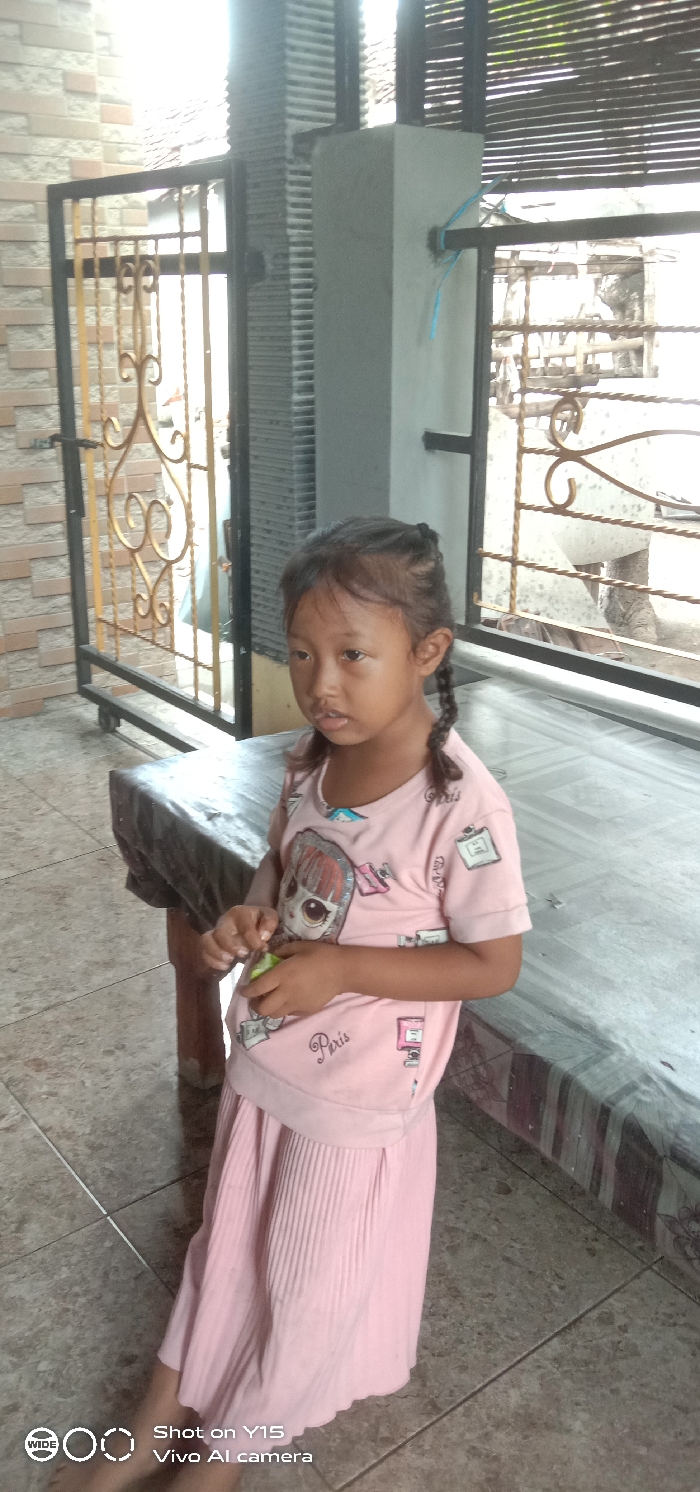 A young female child, approximately 5-8 years old, with dark hair styled in two braids, stands or sits in a semi-outdoor covered area in the city of Sebaung, Indonesia. She is wearing a light pink short-sleeved t-shirt with a large doll graphic and the word "Paris" printed on it, and a matching light pink pleated skirt. Her bare feet are visible at the bottom left of the frame. She holds a small, light green, elongated object in her hands, clasped at waist level. Her gaze is directed towards the left side of the image. The background features a wall with light-colored brick-patterned tiles on the left and a dark-colored metal gate with gold ornamental scrollwork on the right. Beyond the gate, bright daylight illuminates a blurred outdoor area. A concrete pillar is visible behind the child. A dark, rectangular, possibly wooden or tiled, bench or table is present to the child's right. The ground is covered with light-colored square tiles. A light blue cable is tied to a metal bar on the right side of the gate structure.
