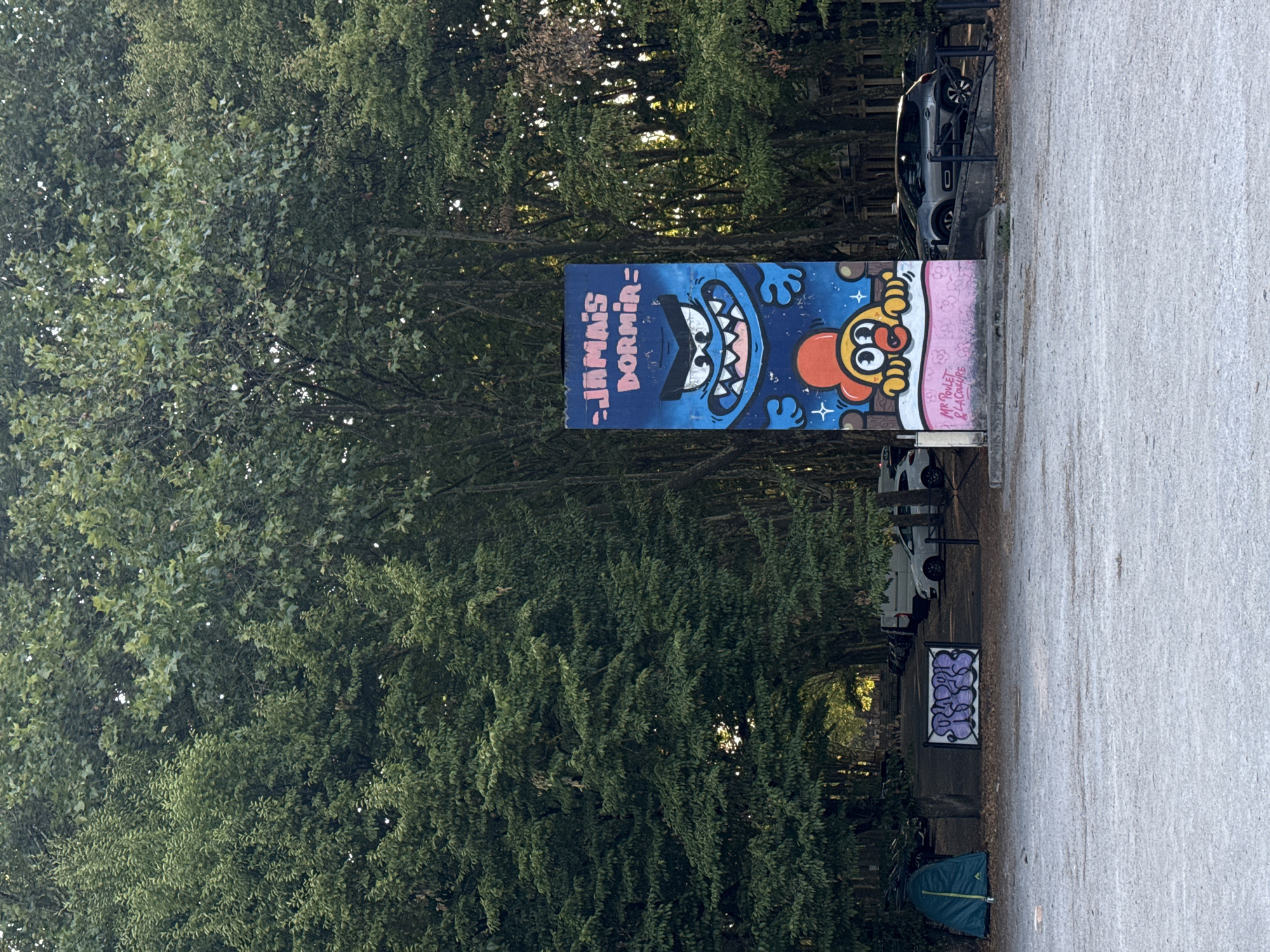 The image depicts a street art mural on a tall, slender structure, likely a utility pole or a custom-built display, in what appears to be an outdoor urban or park setting. The mural is predominantly blue and features a cartoonish monster with a wide, toothy grin and menacing eyes at the top, with the text "JAMAIS DORMIR" written in pink above it. Below the monster, a cheerful, anthropomorphic chicken character is depicted, wearing a red hat and yellow body, lying in a bed of white with pink floral patterns. The bottom of the mural has a signature that reads "MR POULET RLA CULTURE".
The structure the mural is on is a light grey concrete base with a pink band at its bottom. To the left of the mural, a blue tent is pitched on a gravelly ground. In the background, a row of cars are parked under a canopy of mature trees with dense green foliage. The trees form a natural tunnel or walkway, suggesting the location might be a park or a residential area with tree-lined streets. The overall atmosphere is one of an outdoor space with a touch of urban art. The setting suggests it is an open, publicly accessible area, possibly a park or a parking lot adjacent to a wooded area. The lighting suggests it is daytime, with the sun filtering through the trees.