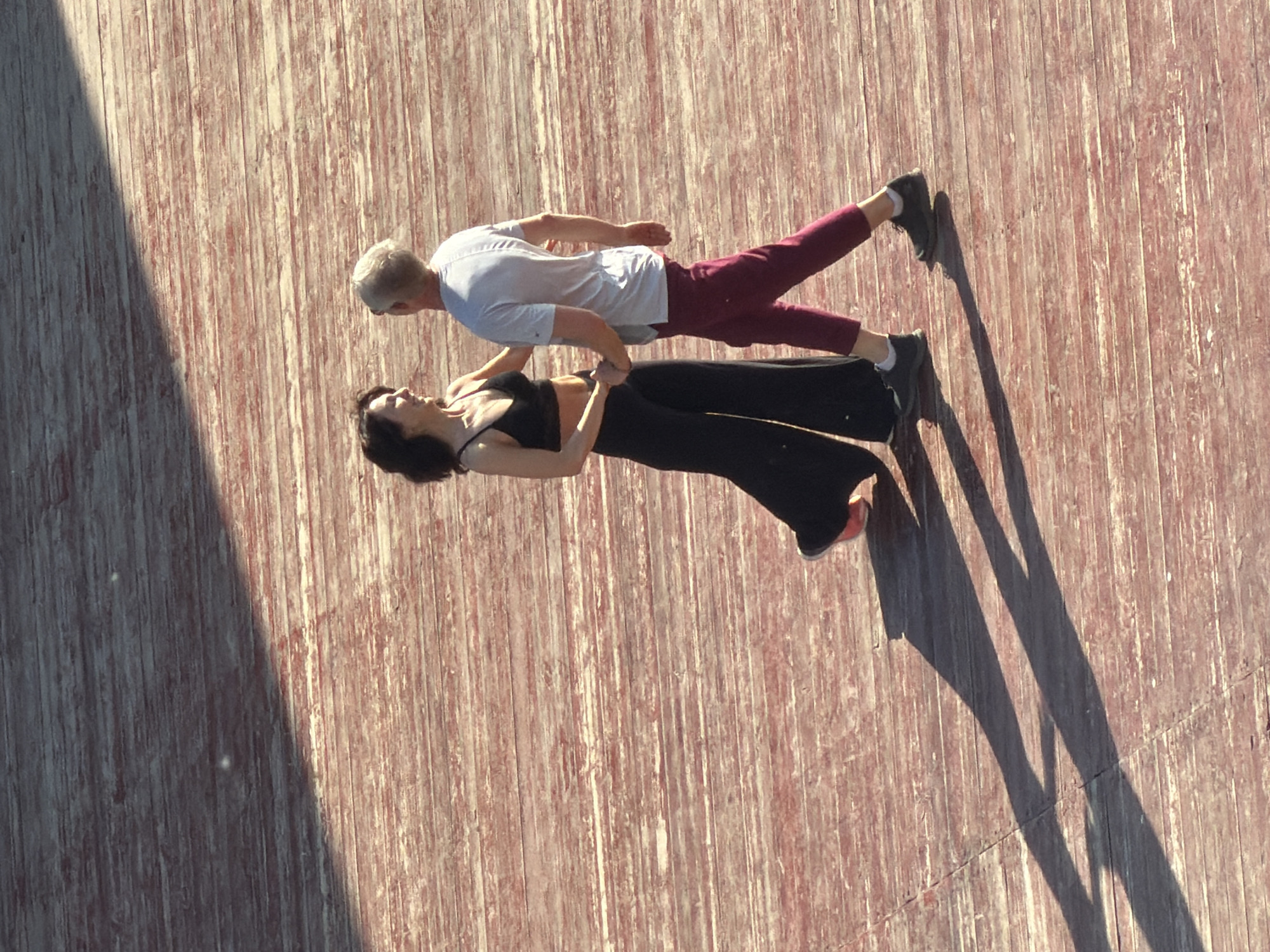 Two people are performing what appears to be a partner acroyoga pose on a wooden surface, possibly a stage or a deck. One person, wearing a black top and black pants, lies supine on the surface. The other person, wearing a white t-shirt and maroon pants, is balancing on the first person's feet and hands, creating a plank-like position. The lighting suggests it is daytime, with shadows cast by the people. The wooden surface appears weathered. The location cannot be definitively determined as Moscow, Russia, from the image.
