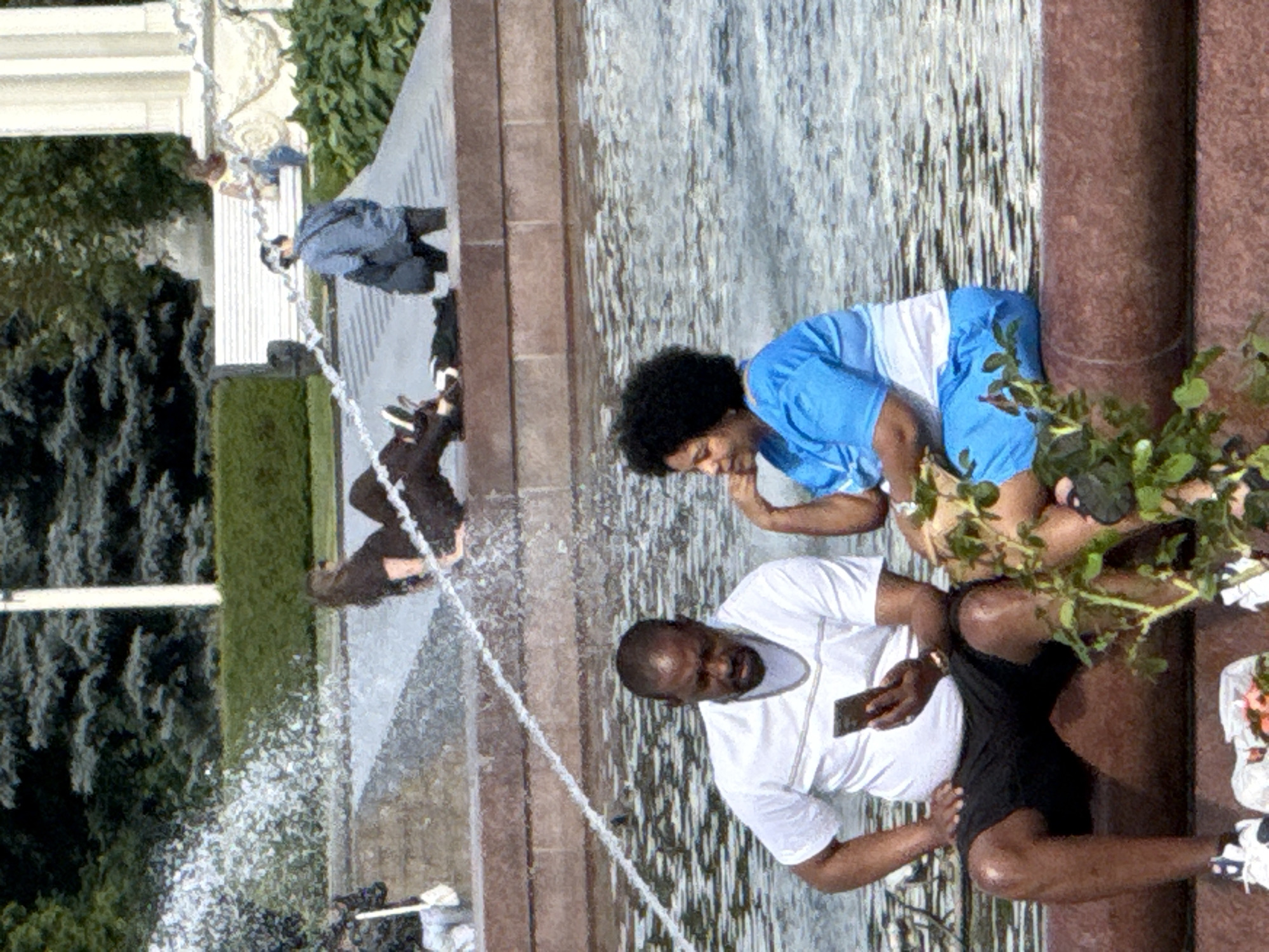 This media file captures a candid moment near a fountain in Moscow, Russia. The scene features at least three individuals engaged in leisurely activity. In the foreground, a man wearing a white t-shirt and black shorts is holding a smartphone and looking towards the camera with a smile. To his left, a person with dark, curly hair is wearing a bright blue top and white shorts, leaning forward and appearing to be interacting with something in the fountain. Further back, a third person in a blue jacket and dark pants is partially visible, looking downwards.
The setting is an outdoor urban environment, likely a park or public square, characterized by a fountain with flowing water and stone structures. Lush green trees are visible in the background, suggesting a pleasant day, possibly in the warmer months. The time of day appears to be daytime, with ample natural light. The overall mood seems relaxed and informal, as individuals are enjoying their time by the water. No text is visible in the image.