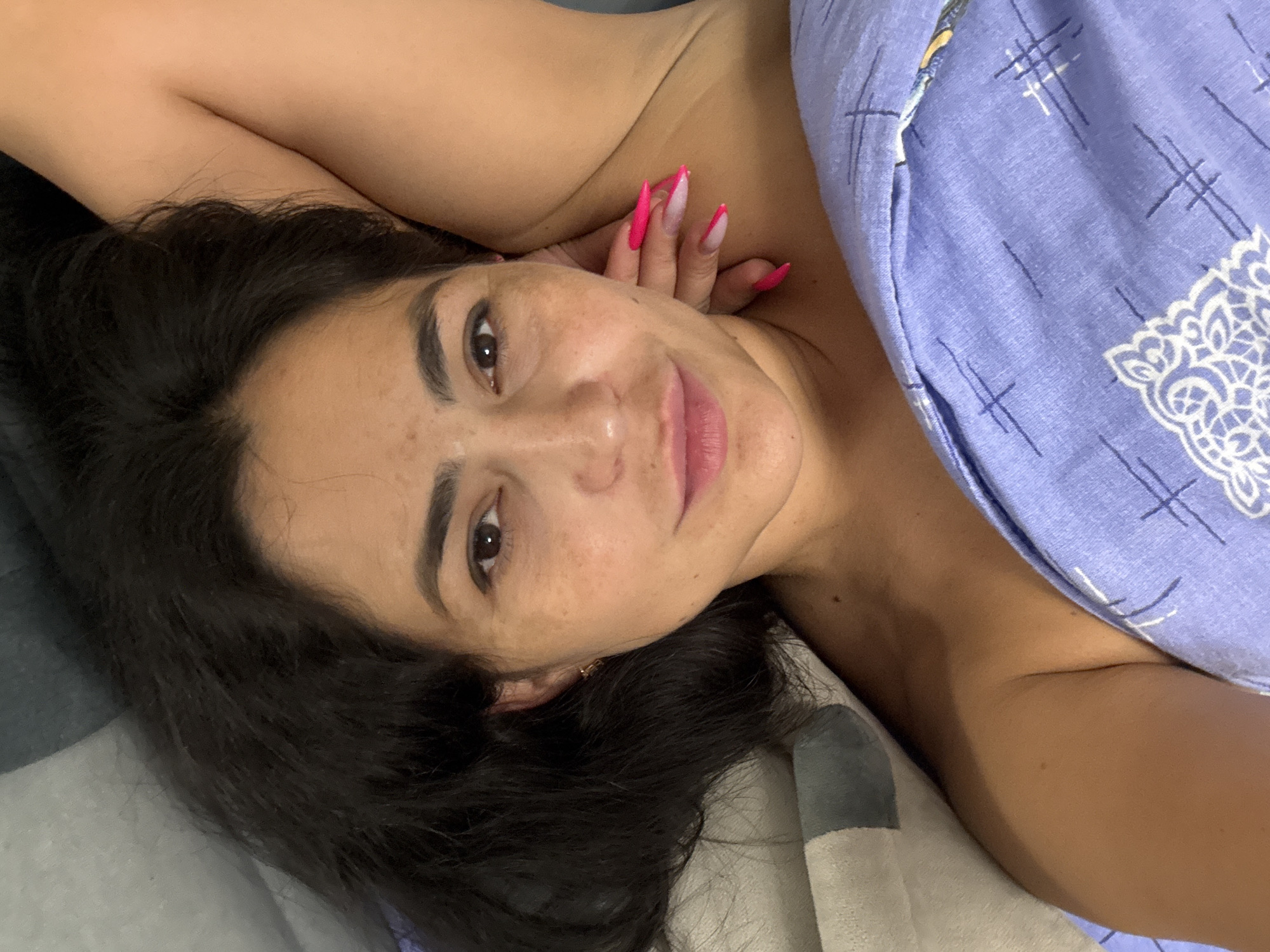 A woman is lying on a bed, partially covered by a light blue blanket with a white geometric pattern. Her head is resting on the bed, and her dark hair is spread around her face. She is looking at the camera with a slight smile. Her right arm is bent, and her hand is near her face, displaying long pink and white manicured nails. The background is a blurred gray and white bed covering. There is no clear indication of the location being Kauchuk Stantsiya, Russia.
