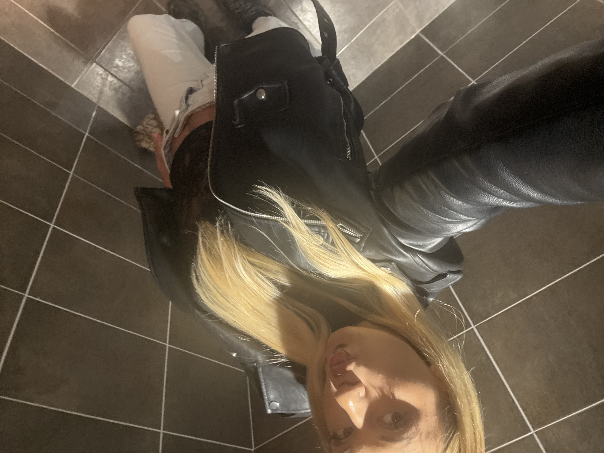 A person is depicted in an indoor selfie, with the image rotated vertically. The subject, appearing to be female, has long, light blonde hair that cascades down her front. She is looking directly at the camera with slightly parted lips. Her face is visible from the nose downwards, and one eye with visible makeup is clearly seen.
She is dressed in a black leather jacket featuring prominent silver zippers and a snap-button pocket on the chest. A dark, possibly lace, garment is visible beneath the open jacket. She is also wearing light-colored trousers or jeans and dark footwear, possibly boots, partially visible at the bottom of the frame. A black bag strap is draped over her shoulder.
The background consists of a dark tiled floor with light grout lines, suggesting an indoor setting. The lighting appears to be artificial, casting some soft shadows. No specific activity beyond posing for the selfie is discernible. There is no visible text in the image. This scene takes place in Stara Zagora, Bulgaria.