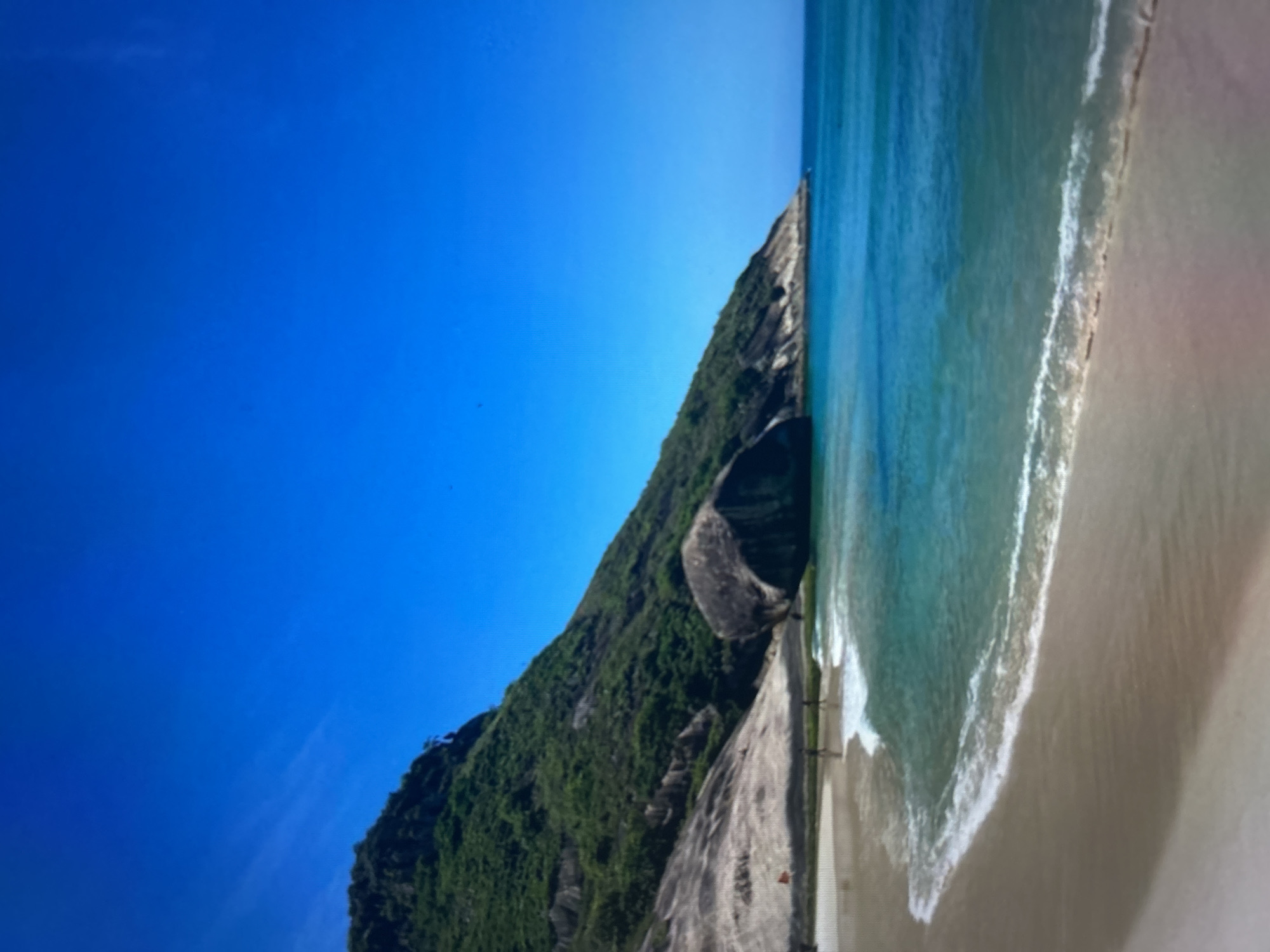 The image depicts a wide, sandy beach meeting a clear, turquoise ocean under a vast, cloudless blue sky. To the left, a steep, green-covered mountain slopes down towards the water. Large, rounded rocks are scattered near the base of the mountain. A few people are visible on the beach, some walking near the water's edge, and a red flag is planted in the sand. The waves are gently breaking on the shore, creating white foam. The scene suggests a peaceful, tropical coastal environment.