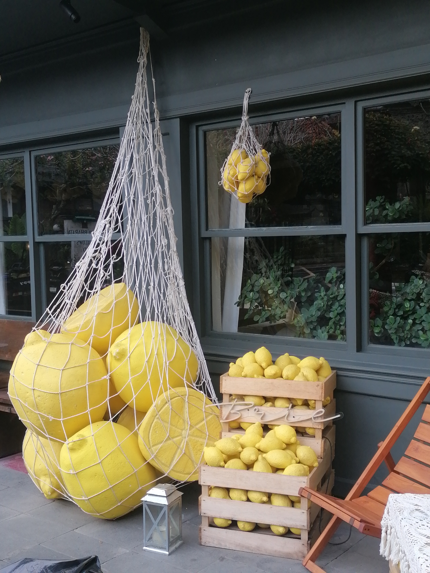 This image depicts an outdoor display of large, yellow, artificial lemons arranged in a net bag and wooden crates. The display is set against a dark green building with large windows that reflect the surroundings. Two windows are visible, one higher and smaller, and another larger one below.
In the foreground, a large white net bag filled with many oversized yellow lemons hangs from the building's overhang. Below this, at ground level, are several wooden crates stacked on top of each other, also filled with yellow lemons. A small, silver lantern with a candle inside sits on the pavement in front of the crates. To the right, a rustic wooden chair with a white textile draped over its arm is positioned next to the crates.
The overall impression is one of a decorative display, possibly for a shop or cafe, with a focus on the vibrant yellow of the lemons. There are no people visible in the image. The setting appears to be a pedestrian area or sidewalk, as suggested by the pavement. The background visible through the windows shows greenery and a reflected image of what appears to be another building.
It is not possible to determine the specific location within Ubud District, Indonesia, from the provided image. The image shows a general outdoor scene with a storefront and decorative elements.
