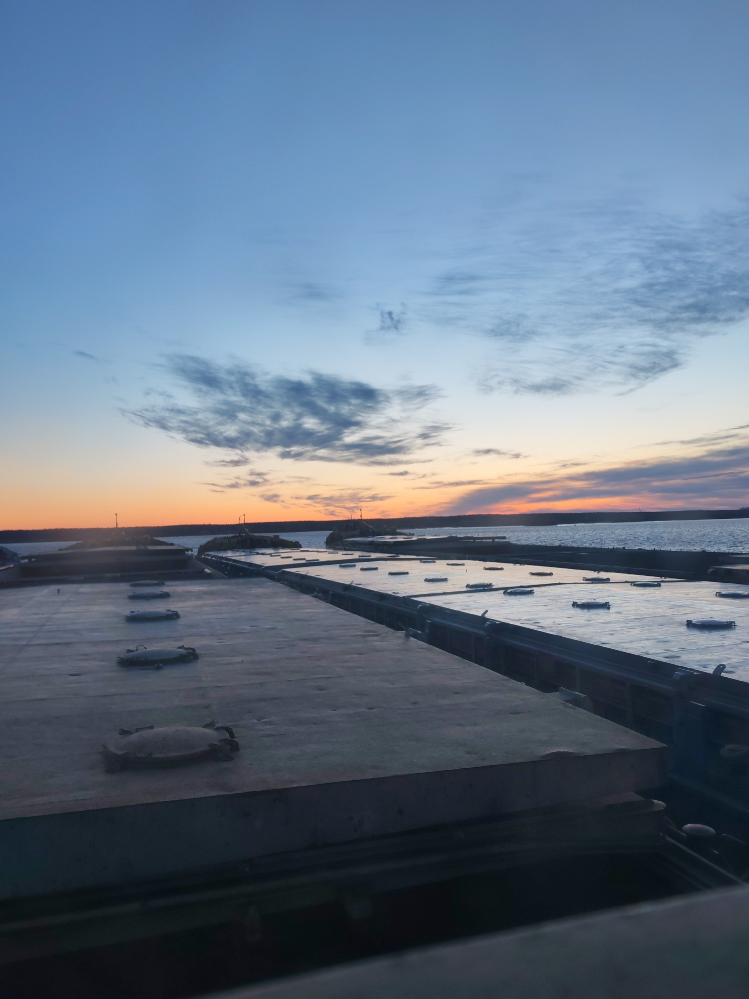The image depicts a view from what appears to be the deck of a ship or large platform, likely near a body of water. The foreground shows a series of rectangular concrete or metal surfaces with hatches or access points. In the background, a body of water stretches to the horizon under a sky transitioning from blue to orange, indicating either sunrise or sunset. The horizon line features a dark landmass or coastline.
While the image is visually similar to scenes found in port areas, there is insufficient information in the image to confirm the precise location as Rybinsk, Russia, or provide any specifics about people, actions, or interactions within the scene. The description is limited to the elements directly visible in the image.