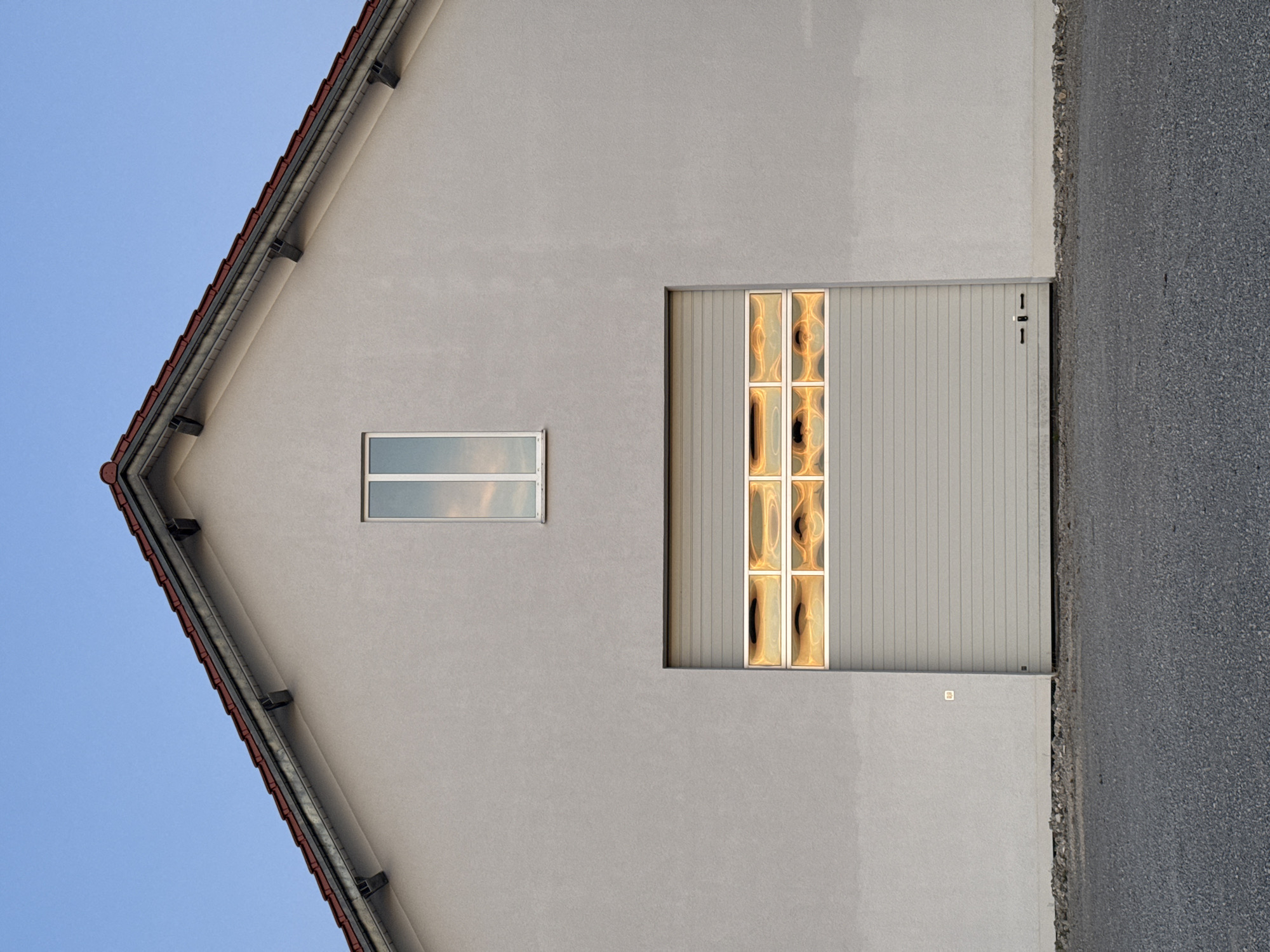 The image shows the facade of a light-colored building with a gray roller garage door. Above the garage door, there is a row of illuminated glass blocks with a warm, orange glow. A narrow, rectangular window with white framing is positioned centrally above the glass blocks. The building has a red-tiled roof with dark gutters. The scene is set against a clear, pale blue sky. In the foreground, a dark asphalt surface leads to the building, with a strip of gravel at the base of the facade. The image was taken in Wettstetten, Germany.