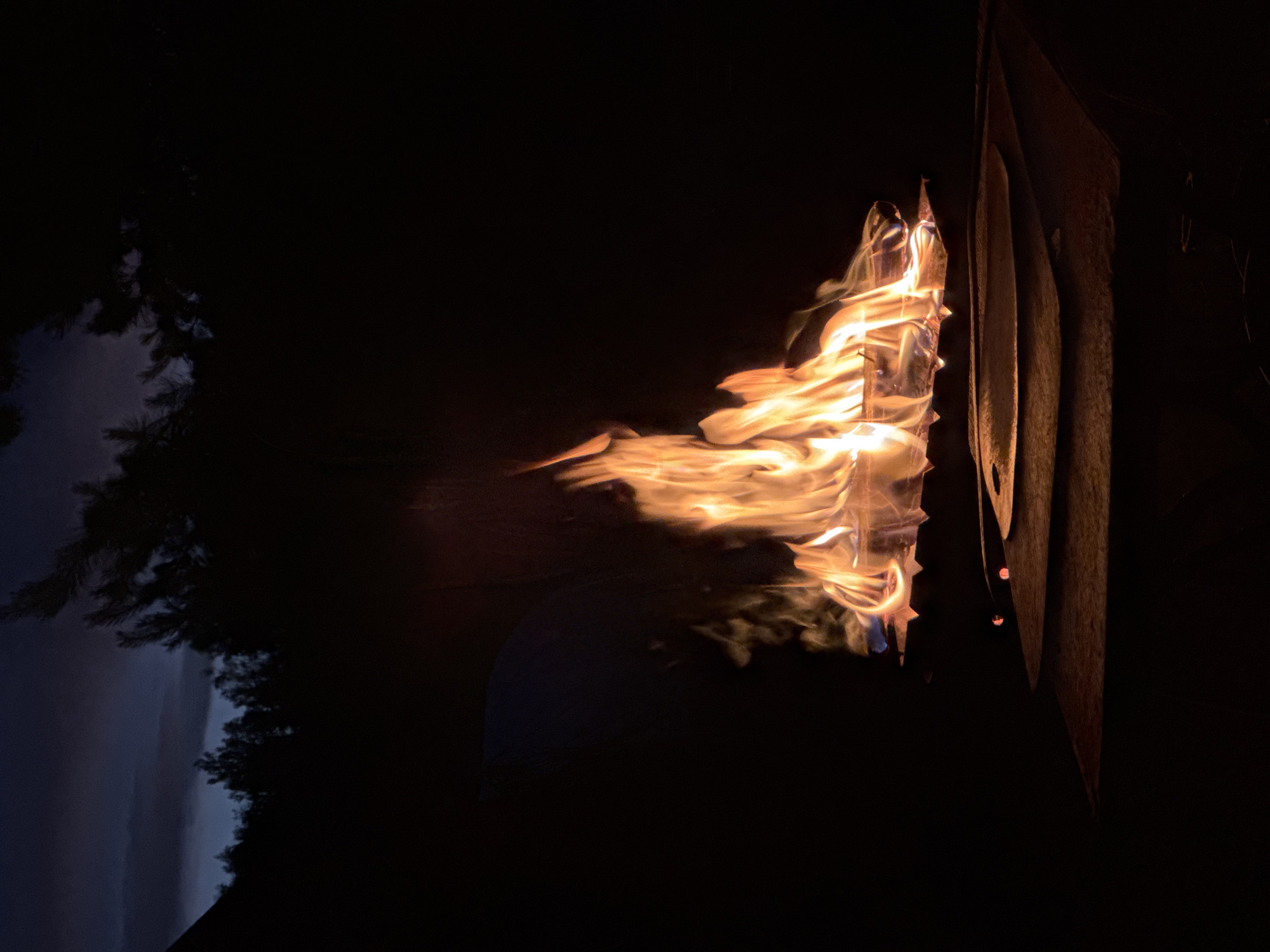 In the city of Поварёнки, Russia, a fire burns in a dark outdoor setting. The flames, a vibrant orange and yellow, rise from a grate holding logs of wood. Below the grate, a circular metal surface with layered, curved edges is visible, casting a shadow on a flat surface beneath it. The background is predominantly black, with the silhouette of trees and a dark, cloudy sky visible at the top of the image. The overall scene is one of a nighttime campfire.