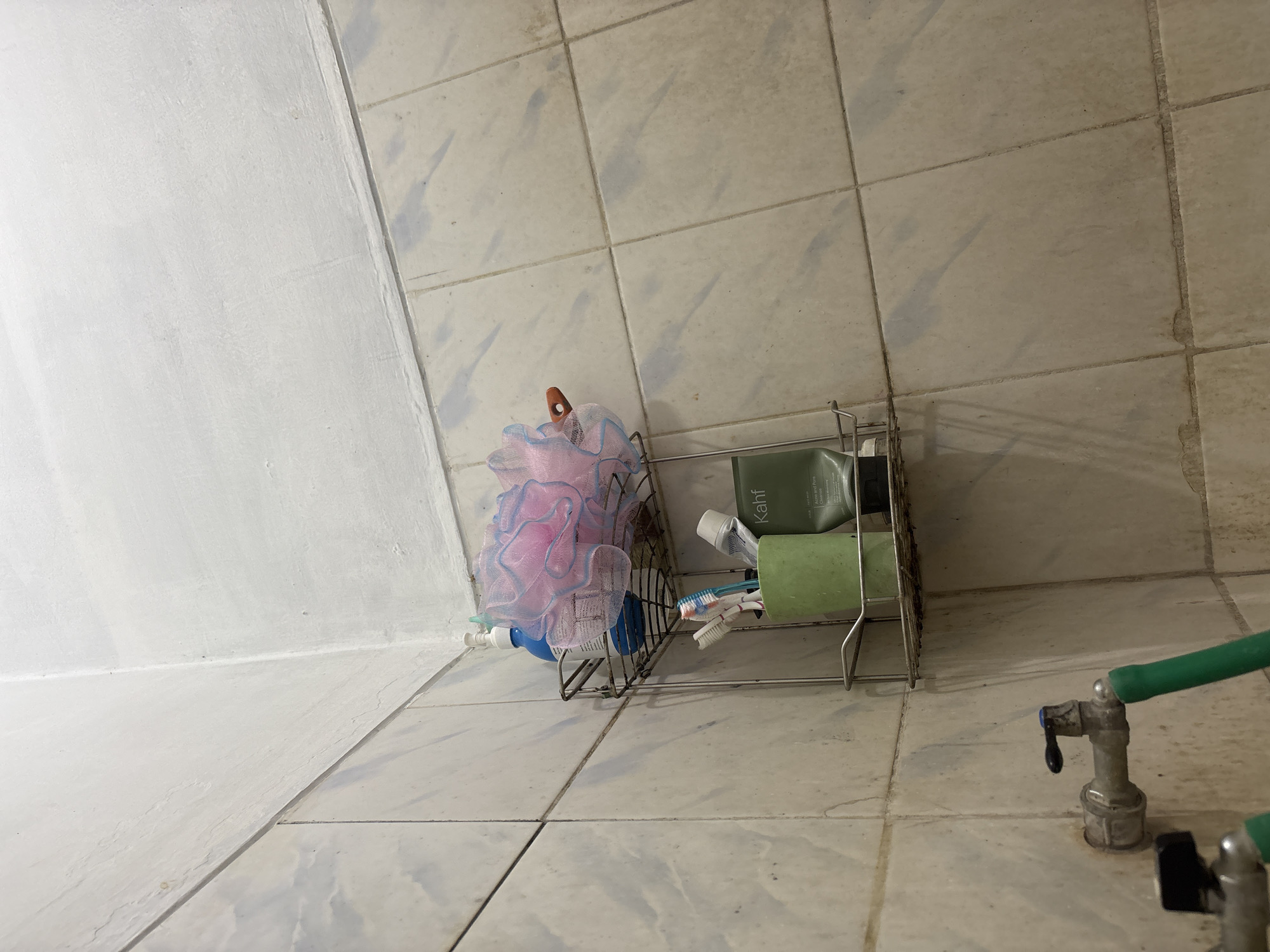 The image depicts a corner of a bathroom, likely in a residential setting in Palimanan, Indonesia. The walls are tiled with a beige, stone-like pattern and the ceiling is painted white, showing signs of wear and tear. A two-tiered metal wire shelf is attached to the wall. The top shelf holds a blue plastic bottle of liquid soap and a pink and blue mesh bath pouf. The bottom shelf contains a green cup holding three toothbrushes, a tube of "Kahf" acne and pore clearing product, and possibly other toiletries. In the lower left of the frame, a metal faucet with a black handle is visible, connected to a green hose. The overall environment appears functional but somewhat neglected, with visible grime on the tiles.