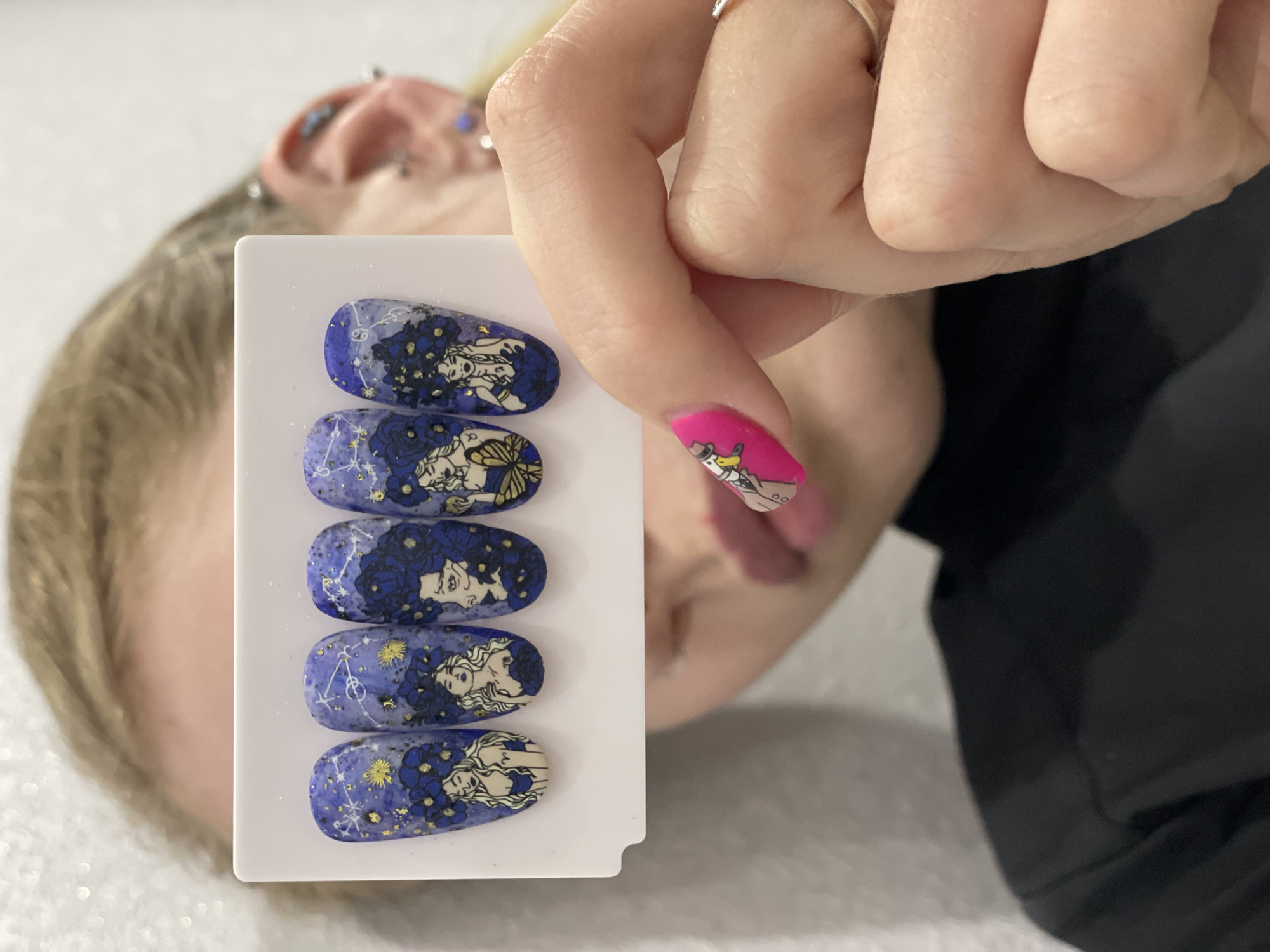 A person lying down holds a white card with five blue and white nail art designs featuring women, flowers, and constellations. The person's hand is visible, with a pink nail featuring a cartoon character. Multiple piercings are visible on the ear of the person lying down. The background includes a white surface and a black garment. The location is potentially Izhevsk, Russia, though no specific location markers are discernable from the image.