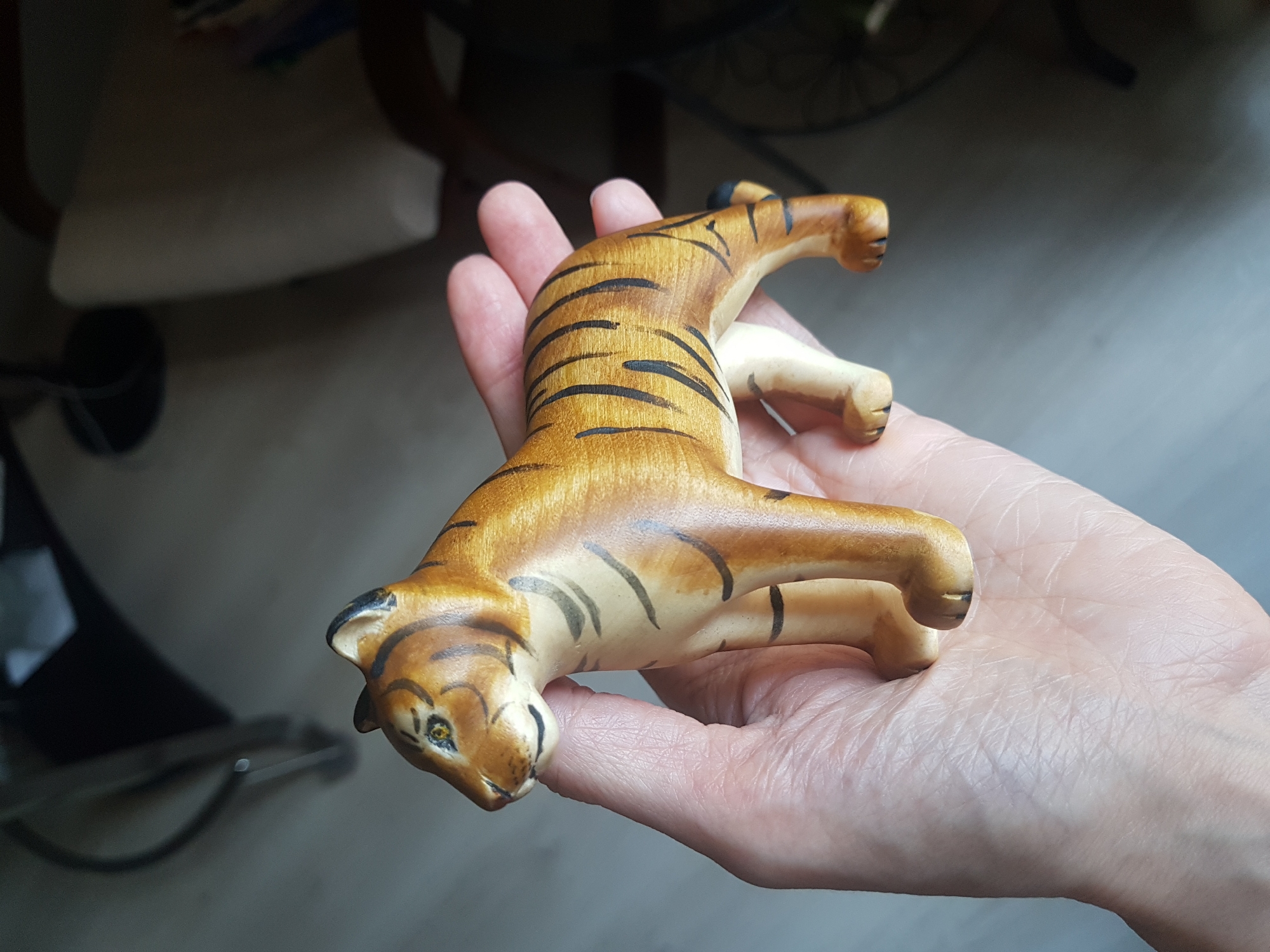 A human hand holds a small, ceramic tiger figurine. The tiger is painted in shades of brown with black stripes, and its legs are painted a lighter beige color. The tiger appears to be posed mid-stride with its head tilted downward. The hand appears to be resting indoors; in the background, there are pieces of furniture, including a table, a chair, and the wheel of a bicycle. There is no evidence to suggest that this image was taken in Saint Petersburg, Russia.