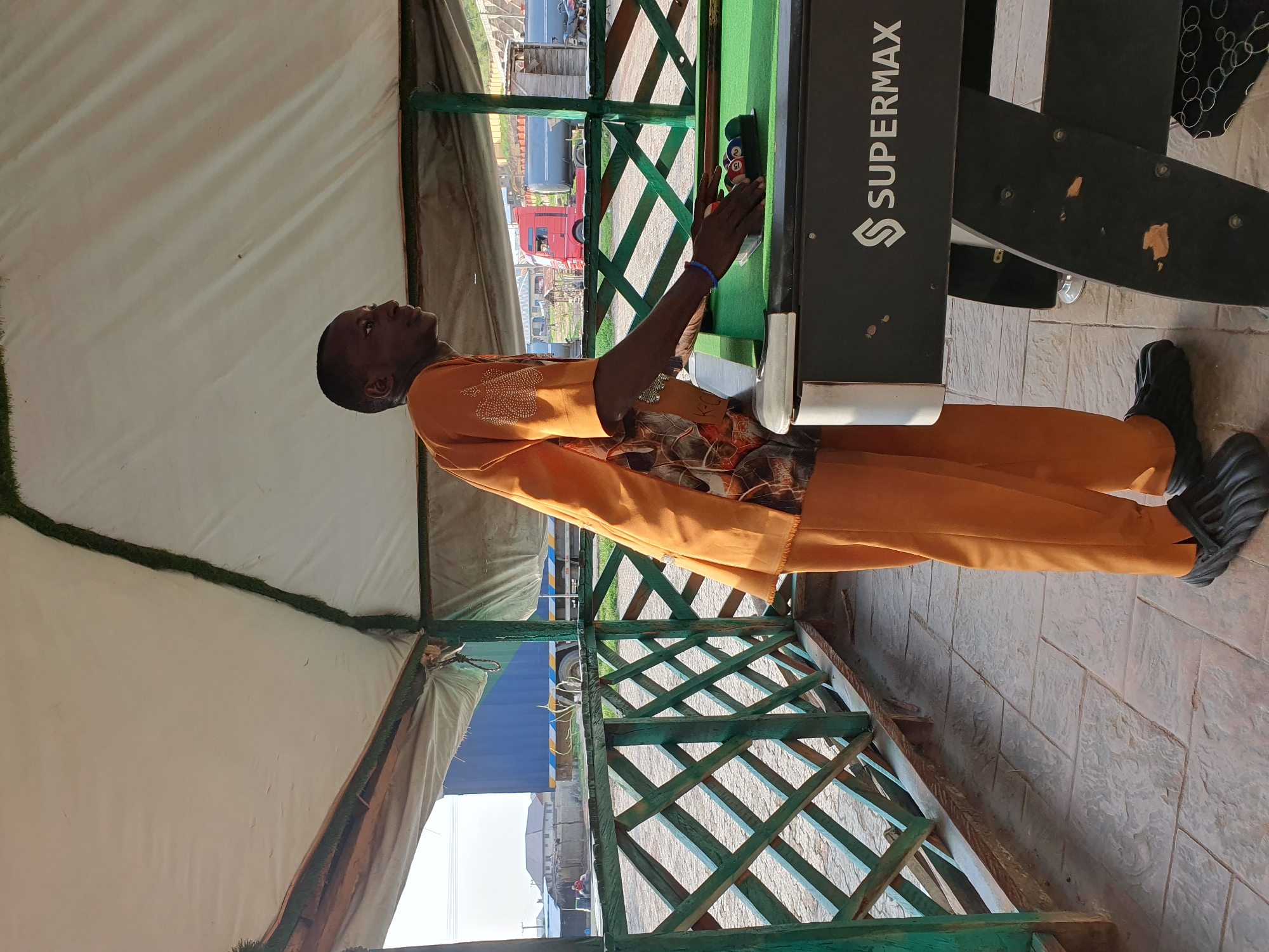 A man in an orange traditional outfit is standing next to a billiard table. The man is looking towards the billiard table, and his hands are positioned as if he is about to play. The billiard balls are racked at one end of the table. The scene appears to be outdoors, possibly under a canopy or tent structure, with a view of trucks and buildings in the background. The floor is tiled. The text "SUPERMAX" is visible on the side of the billiard table. The image seems to have been taken during the daytime.