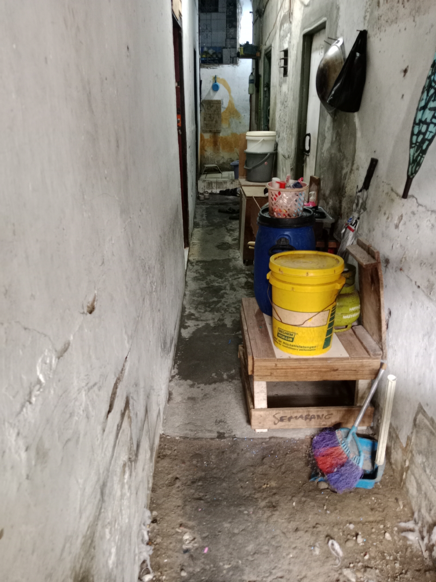 The image depicts a narrow alleyway between two concrete buildings, likely in a densely populated urban area of North Jakarta, Indonesia. The alley floor is made of concrete and appears worn and dirty, with scattered debris.
On the right side of the alley, a simple wooden bench is placed against the building wall. On the bench, there is a large yellow plastic bucket with a lid, labeled with some text, and behind it, a smaller green object that resembles a gas cylinder. A blue barrel is also visible.
Further down the alley, there are stacked white and grey buckets. Above the bench, a dark bag is hung on the wall, next to a door. The walls of the buildings are unpainted concrete, showing signs of age and wear.
In the foreground, on the ground next to the bench, a broom and dustpan are placed. The broom has colorful bristles of red, purple, and blue. Some of the text "SEMARANG" is visible on the wooden structure supporting the bench. The overall impression is of a utilitarian and somewhat neglected outdoor space.