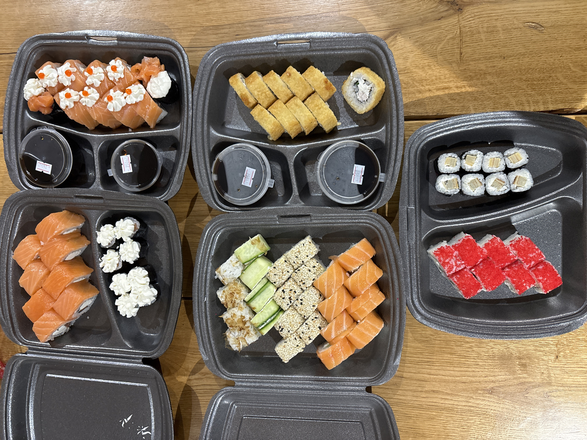 An overhead shot displays an assortment of sushi and rolls laid out in several gray, compartmentalized takeout containers on a light brown wooden surface. Five separate containers are visible, each filled with different types of Japanese-inspired cuisine.
In the top left container, there are eight salmon-wrapped rolls, each topped with a dollop of white cream and a few pieces of orange fish roe. Beside them, a small portion of thinly sliced ginger and another piece of salmon-topped sushi are present. Two small, clear plastic cups filled with dark soy sauce are also in this container, one bearing a small white label with "27092" written in black, and the other with "2808".
The top middle container holds nine pieces of fried or tempura-style rolls, light golden brown in color, with one additional roll visible, showing a white filling. This container also includes two soy sauce cups, one labeled "2708" and the other "2808".
To the top right, a smaller container features eight pieces of maki rolls, which appear to have a light-colored filling, possibly a yellow omelet or cream cheese, and are wrapped in white rice.
The bottom left container showcases ten pieces of salmon-wrapped rolls with a white filling, accompanied by six pieces of dark-wrapped rolls topped with white cream arranged in a decorative, flower-like pattern.
In the bottom middle container, three distinct rows of rolls are presented: one row of five rolls topped with thin cucumber slices, another row of five rolls coated in sesame seeds, and a third row of seven salmon-wrapped rolls.
The bottom right shows six pieces of rolls generously covered in bright red fish roe (tobiko or masago).
The entire setup suggests a takeout or delivery meal, possibly for a group, indicating an event focused on dining. The handwritten numbers on the sauce containers could be order identifiers or production dates. The scene is indoors, with no specific time of day discernible beyond typical meal times.