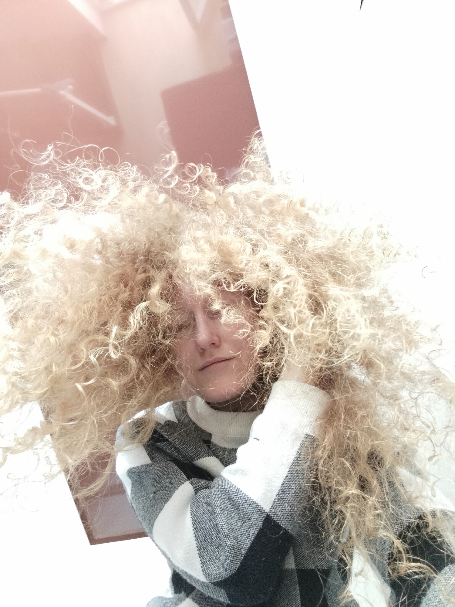 A woman with voluminous, tightly curled blonde hair occupies the majority of the frame, obscuring much of her face except for her nose and mouth. She is wearing a gray and white patterned sweater with a high neckline. The background consists of blurry shapes in shades of white, beige, and peach, possibly suggesting a room interior. There is no readily identifiable information that directly identifies the city of Krykhivtsi, Ukraine. The scene is brightly lit, giving the image a slightly washed-out quality. There are no evident actions or interactions taking place.