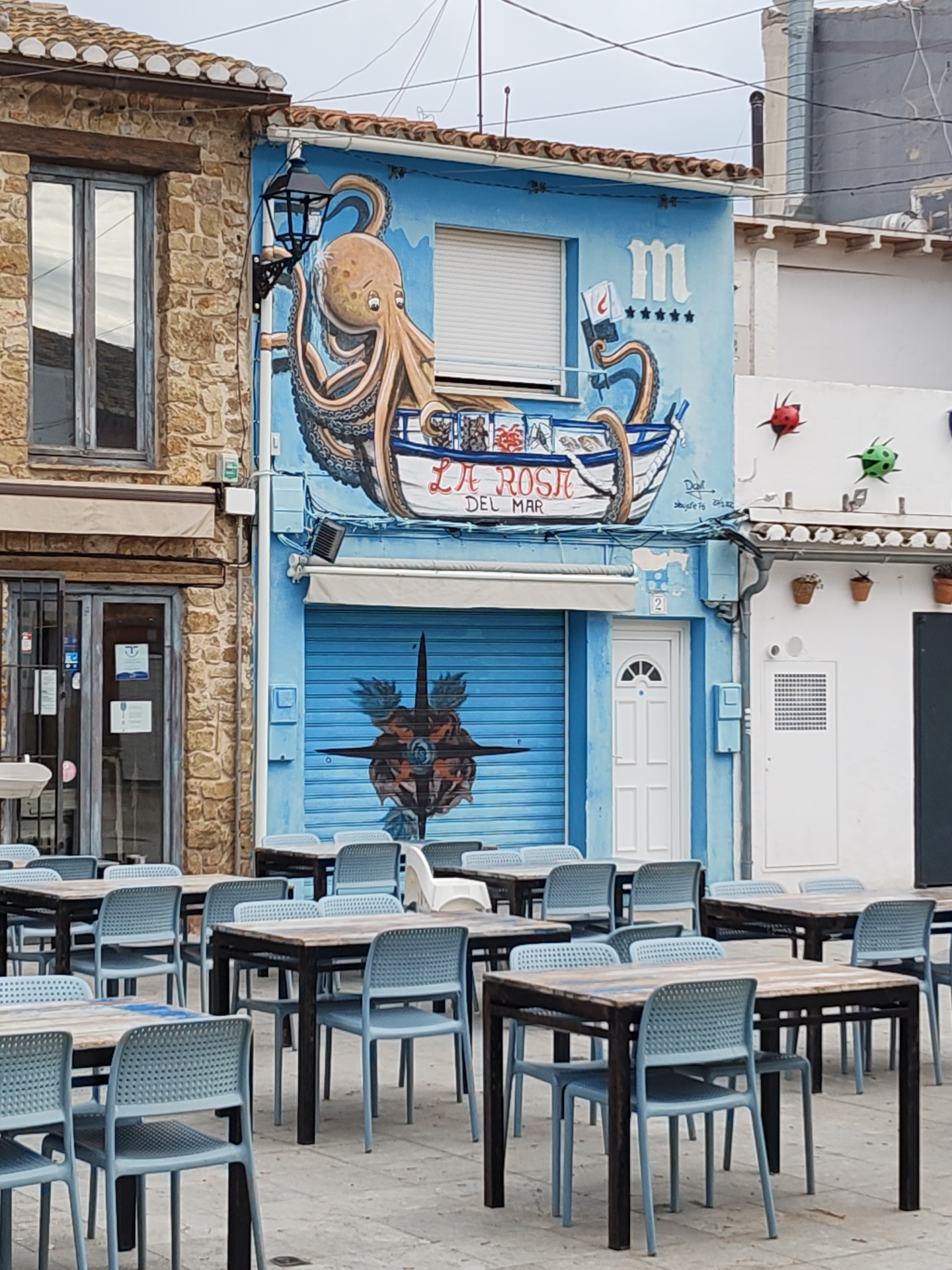 The image shows an outdoor seating area in front of a building in Dénia, Spain. The building is decorated with a mural featuring an octopus in a boat labeled "LA ROSA DEL MAR." A blue garage door is painted with a compass design, and a white door is adjacent to it. Tables and chairs are arranged in the outdoor area. The surrounding buildings are made of stone and stucco. The sky is overcast.