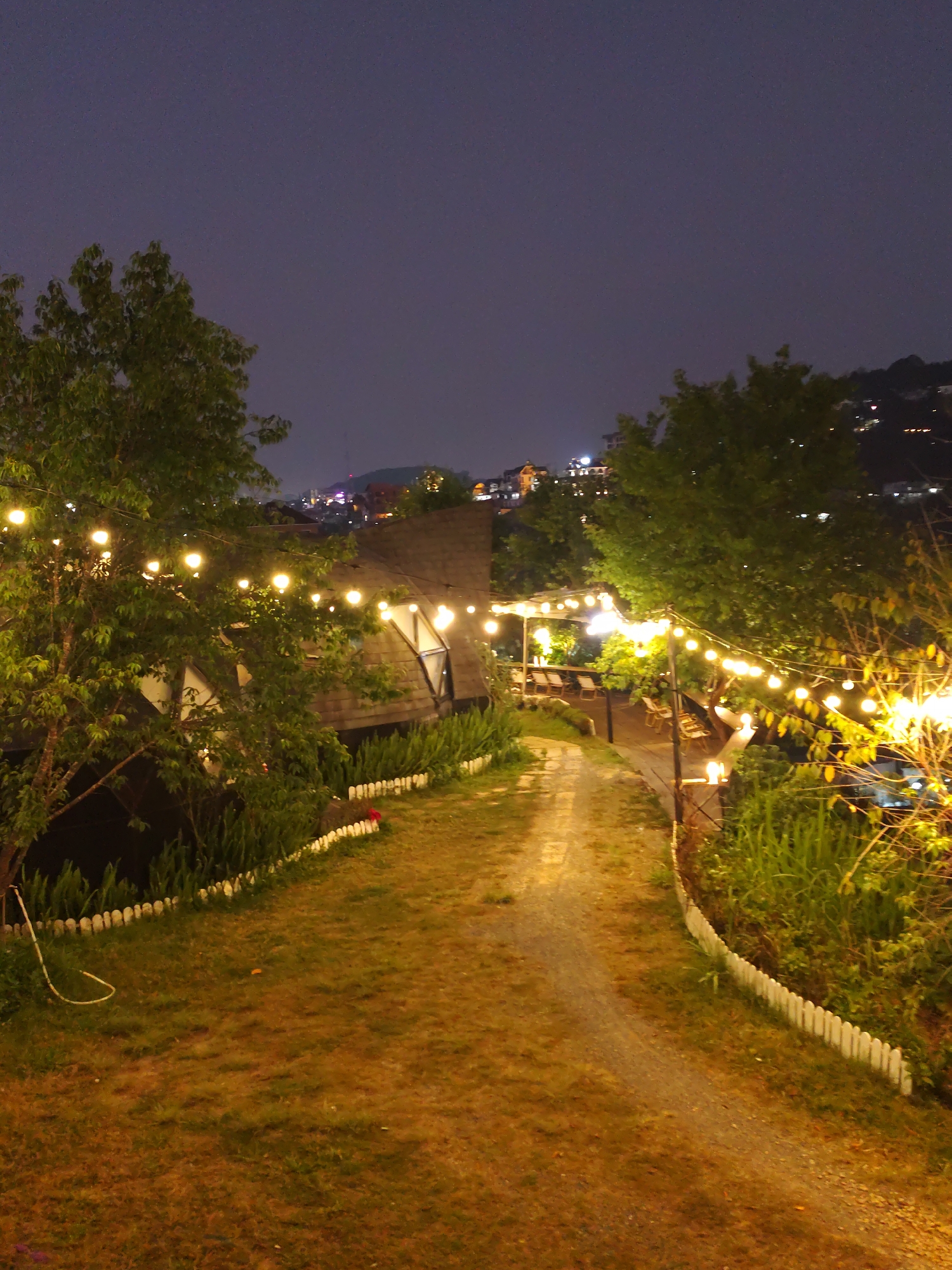 At night, a slightly curving pathway leads through a grassy yard towards a building in Da Lat, Vietnam. Trees frame either side of the path, adorned with strings of illuminated spherical lights. A low, white picket fence borders the grass on both sides. Beyond the building, more lights and structures suggest a townscape situated on a hillside. The sky is dark.