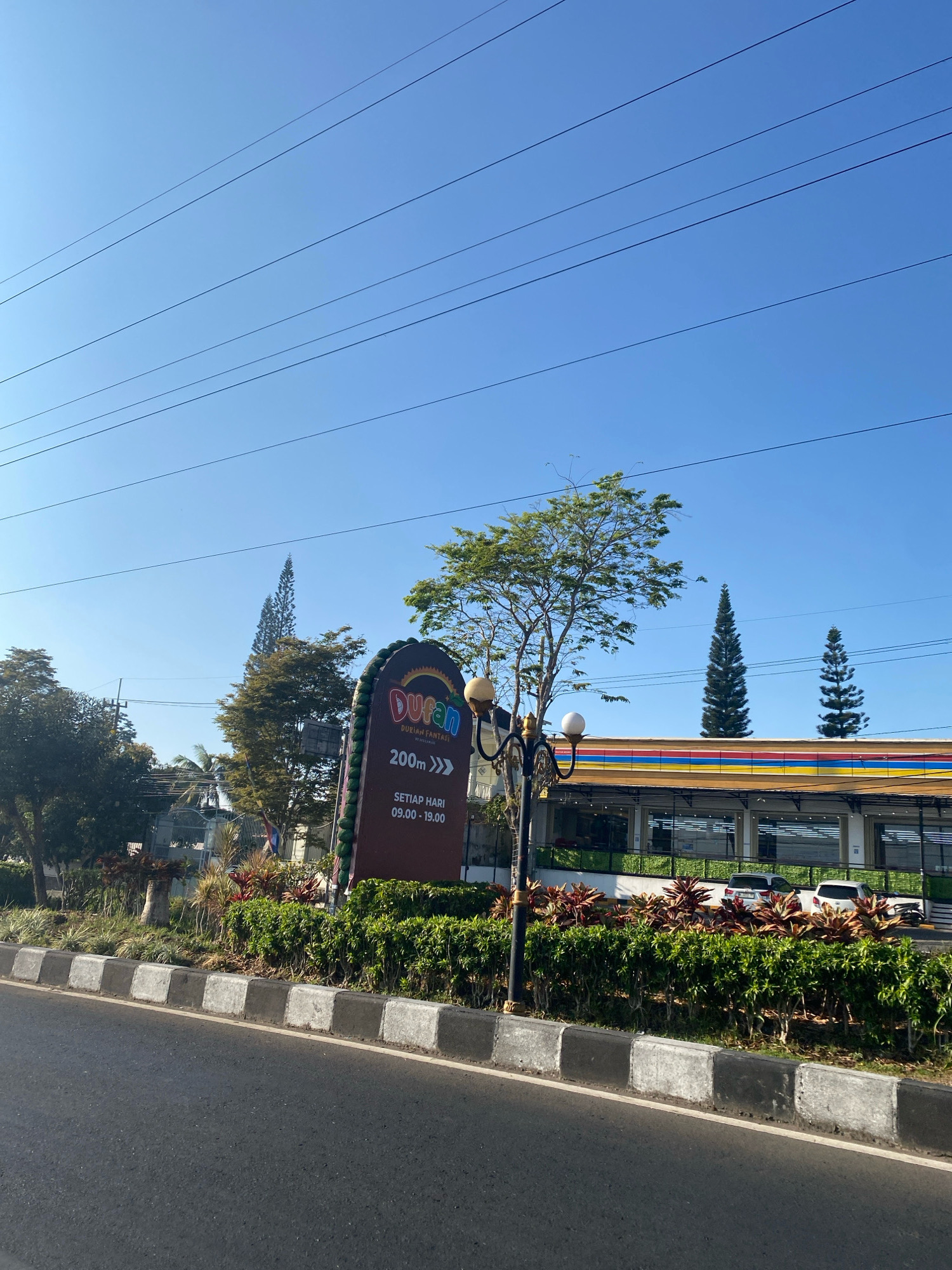 A low-angle, eye-level shot captures a clear, blue sky with overhead power lines crisscrossing the frame. Below the sky, a row of tall, dark green pine trees stands against the horizon. In the middle ground, a building with a colorful facade, featuring bands of yellow, red, and blue, is visible. The building has large glass windows reflecting the surroundings and a covered walkway or awning in front.
To the left of the building, a large, dark brown sign with a colorful "DURAN" logo at the top is prominently displayed. The sign indicates "DURIAN FANTASI" and provides directional information "200m >>>". Below this, it states "SETIAP HARI 09.00 - 19.00," which translates to "Every Day 09:00 - 19:00". A decorative street lamp with two globes stands next to the sign.
The foreground features a paved road and a sidewalk bordered by a curb with alternating black and white concrete blocks. A lush green hedge and ornamental plants with red and green foliage are planted between the sidewalk and the building. Two cars, one silver and one white pickup truck, are parked in front of the building. The scene is bathed in sunlight, suggesting a daytime setting.