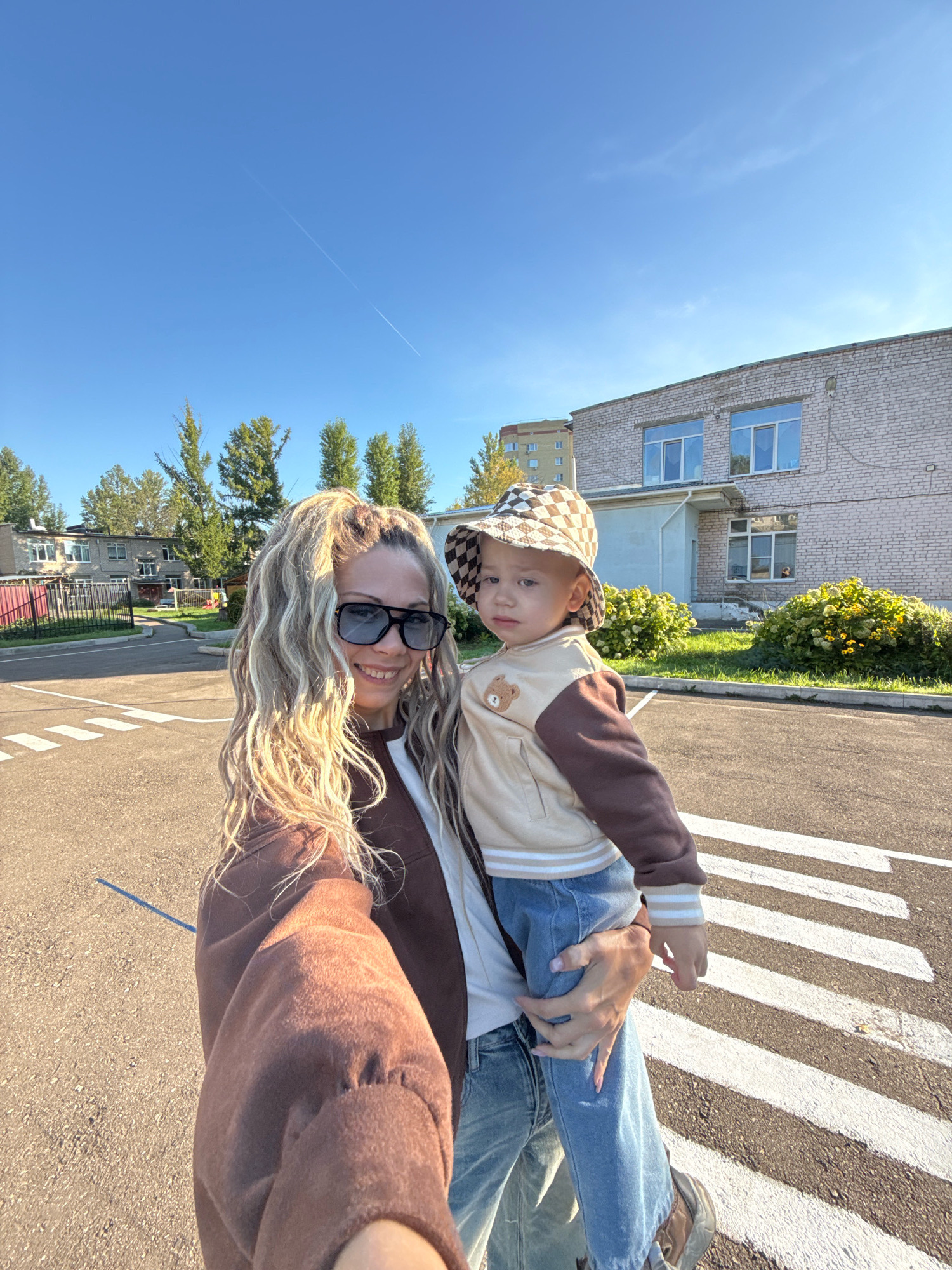 In Rusanovo, Russia, under a clear blue sky with a faint contrail, a woman with long, blonde, wavy hair and sunglasses takes a selfie. She is holding a young child in her arms. The child is wearing a checkered hat and a light brown and white jacket with a teddy bear emblem. They are standing on a paved road with a pedestrian crossing in the foreground. Behind them, a two-story brick building with windows and an entrance is visible, along with green bushes. Further in the background, trees and a taller building can be seen. The overall scene depicts a sunny day outdoors.