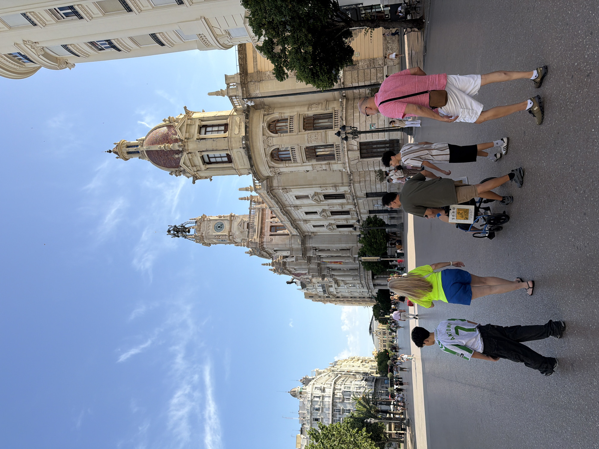 In an outdoor urban square with a wide, paved walkway, several people are walking and standing under a bright blue sky with scattered white clouds. The architecture in the background features ornate buildings with classical influences, including columns, arched windows, and a domed structure topped with a spire. A prominent clock tower rises above one of the buildings. In the foreground, a boy in a green and white soccer jersey with the name "ANTONY" and the number "7" on the back, wearing dark pants, walks away from the camera. To his right, a woman with long blonde hair, wearing a neon yellow top and blue shorts, walks ahead. Several other individuals are visible in the midground and background, including a man in a pink shirt and white shorts, a person in a wheelchair being pushed by another individual, and a group of people on the walkway further away. Trees line the sides of the square, and streetlights are present along the buildings.