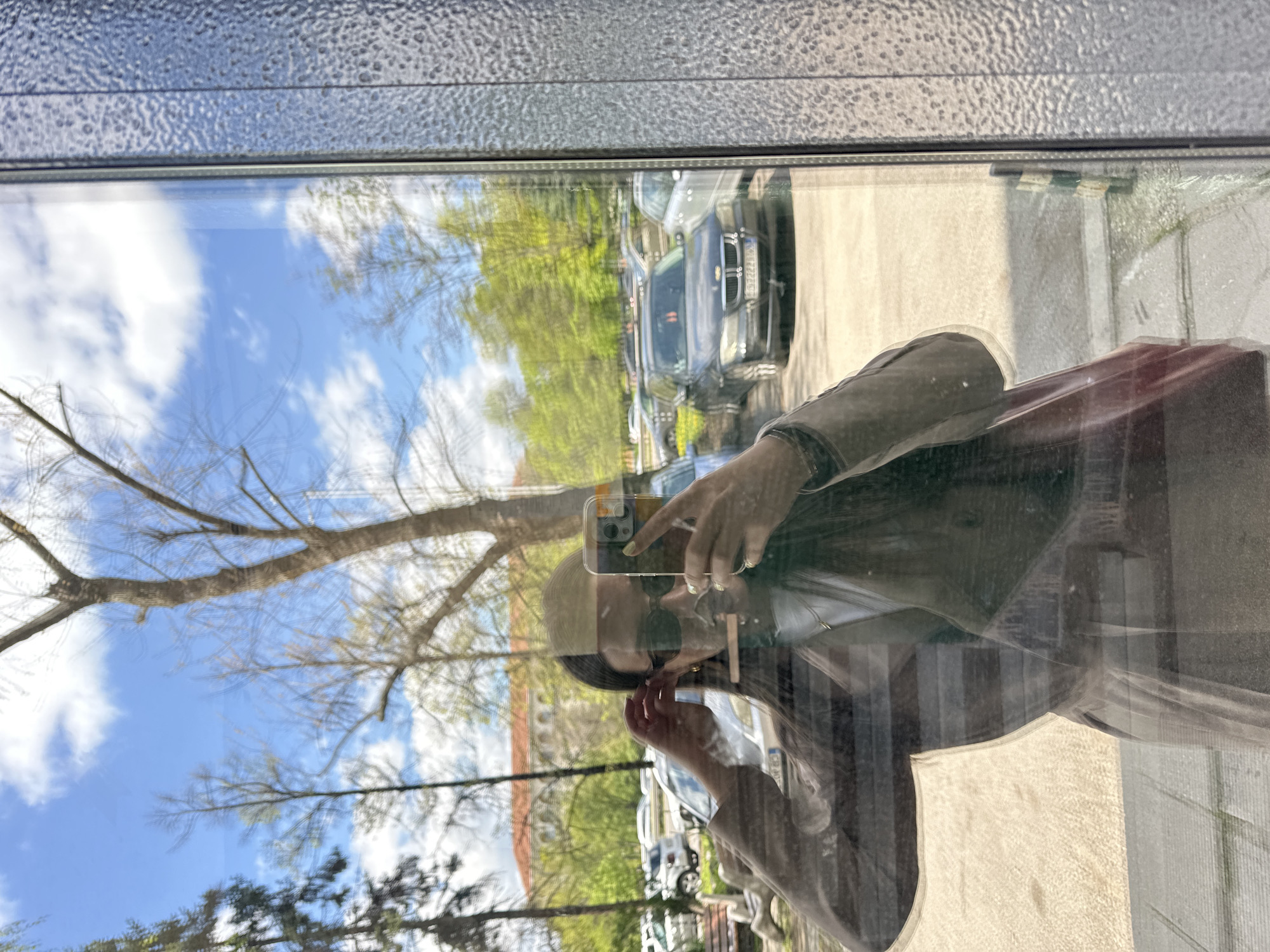 A person takes a selfie in a window. The reflection reveals a streetscape, including a tree, parked cars, and a building with a red roof. The location is suggested to be Sofia, Bulgaria, given the style of architecture.