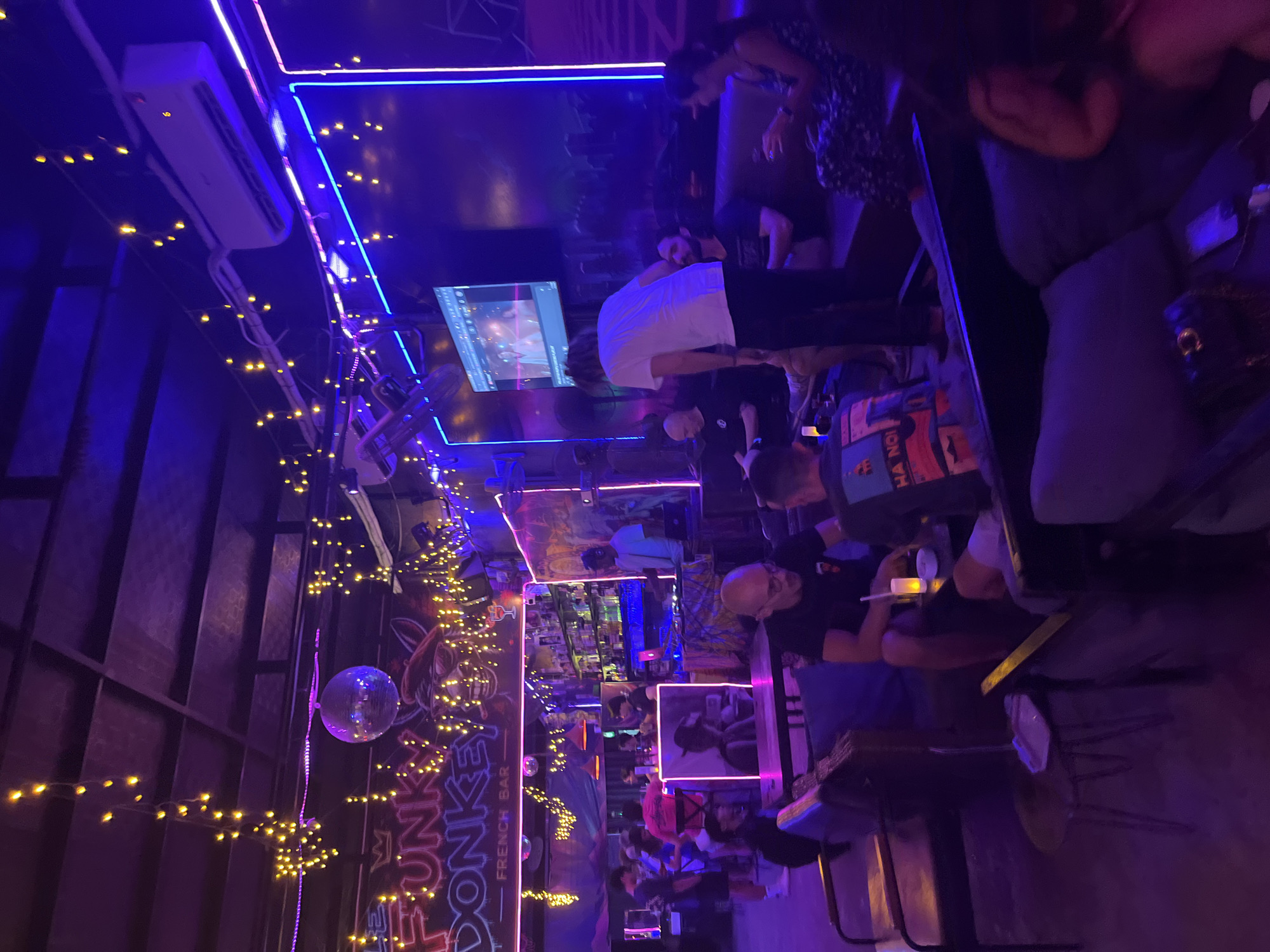 The image depicts the interior of "The Funky Donkey — French Bar" in Đà Nẵng, Vietnam, at night. The space is illuminated by prominent purple and blue neon lighting along the walls and fixtures, complemented by numerous strands of warm yellow string lights draped across the ceiling. A disco ball hangs centrally in the ceiling area.
In the background, a bar counter is visible on the left, stocked with bottles. Adjacent to it, a DJ booth is set up, where a male individual is operating a DJ controller. Several patrons are standing near the bar. A large neon sign reading "THE FUNKY DONKEY — FRENCH BAR" is prominently displayed above the bar area, featuring pink lettering and a stylized donkey logo.
In the mid-ground and foreground, various groups of people are seated on dark upholstered sofas and wooden armchairs, arranged around small tables. Approximately 15-20 individuals, appearing to be adults, are present. Patrons are engaged in conversation, holding drinks, and observing the environment. One person in a white t-shirt stands near a table in the center, facing away from the camera, interacting with seated individuals. A man with glasses, wearing a black t-shirt, is seated in the foreground on the left, holding a glass. On the far right, a woman in a patterned dress is seated on a sofa, holding a drink. Another person in the foreground wears a shirt with "HA NOI" printed on the back.
A flat-screen television mounted on the right wall displays a video. Ceiling-mounted air conditioning units and fans are also visible. Small tables hold items such as drinks, glasses, and a lit candle.