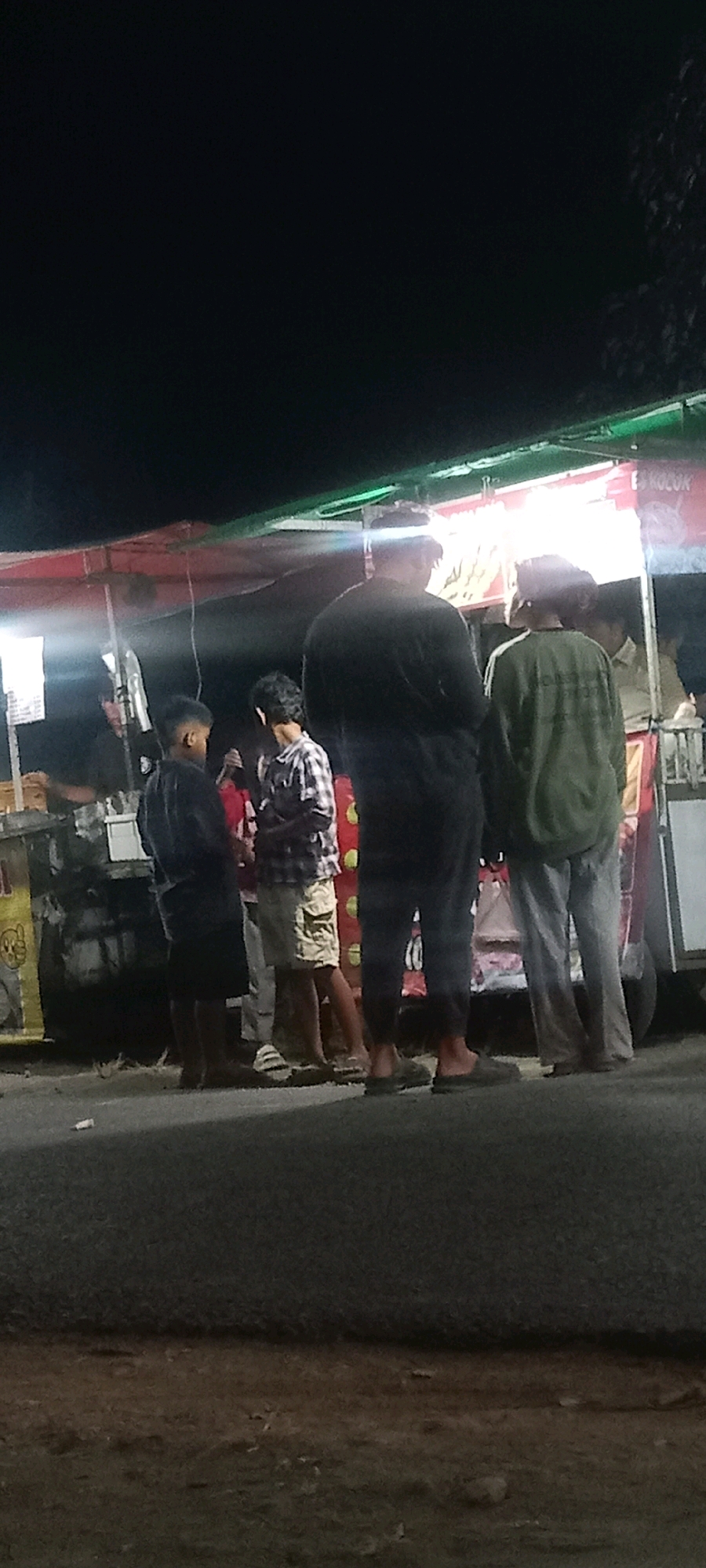 The image depicts a nighttime scene at a food stall in Kras, Indonesia. Four individuals are visible, primarily from the waist down. In the foreground, two individuals stand close together. The person on the left wears a black shirt and dark shorts, while the person on the right has on a checkered shirt and light-colored shorts. Further back and to the right, two taller individuals are observed. The one in the center wears a dark long-sleeved shirt and dark pants, and the individual on the far right is in a green long-sleeved shirt and light-colored pants. They appear to be standing in front of a brightly lit food stall, with some of the stall's structure and signage partially visible. The ground in the foreground is a paved surface, likely a road or sidewalk, with some debris scattered. The overall ambiance suggests a casual street-side food vendor operation.