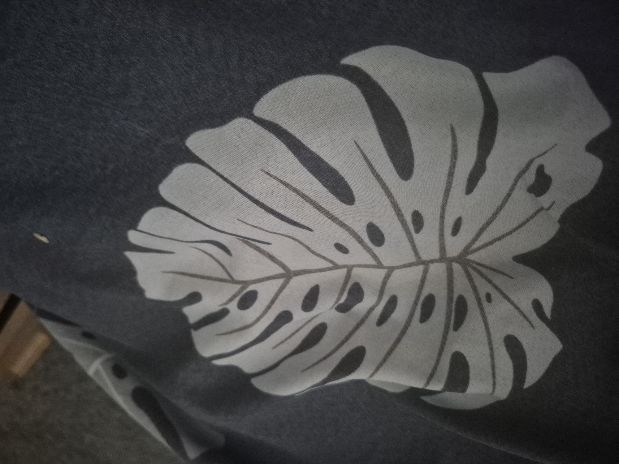 The image displays a close-up view of a dark gray textile patterned with a large, stylized Monstera leaf. The leaf design is light gray or off-white, featuring distinct veins and characteristic fenestrations (holes) rendered in a darker gray or black. The background fabric is a solid dark charcoal or slate gray, exhibiting a subtle woven texture. The material appears soft and shows some natural draping or creasing, suggesting it could be part of clothing, bedding, or a decorative item. A small, faint yellowish speck is visible on the dark fabric above and to the left of the leaf pattern. In the lower left corner, a blurred, light brown object, possibly a piece of wooden furniture, is partially visible, indicating an indoor setting. There are no people present, and no specific activity or event is taking place. No text is visible in the image.