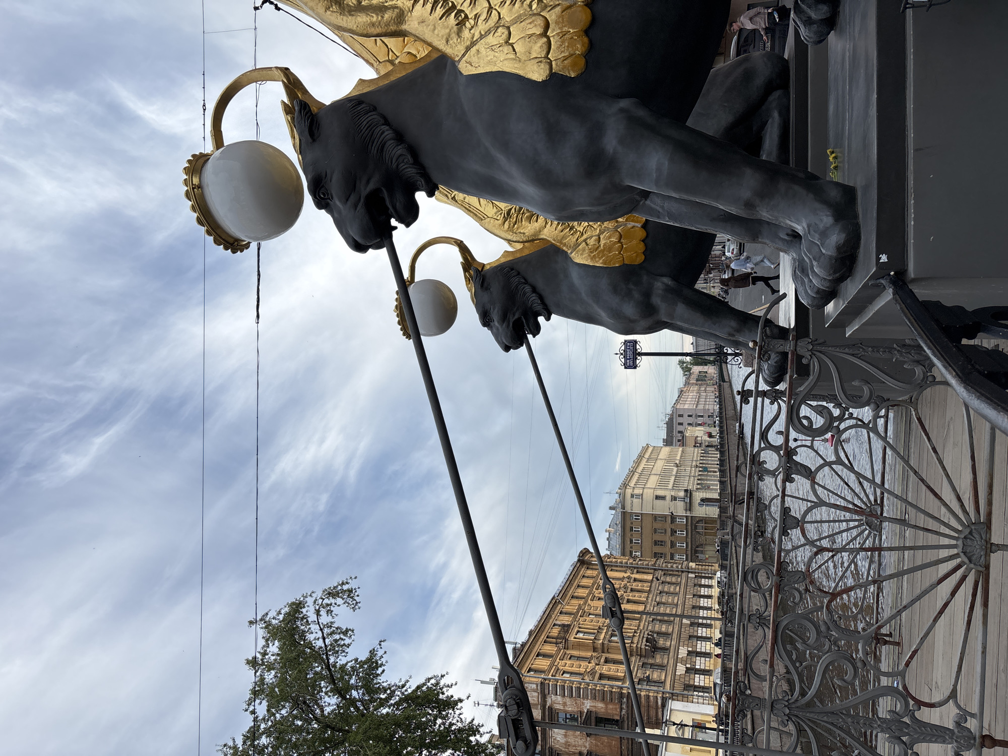 The image, when viewed upright, prominently features two large, dark-colored statues of winged griffins with golden wings. Each griffin holds a lamppost in its mouth, from which a spherical white light fixture hangs. The griffins are detailed with strong, powerful forms, and their golden wings shimmer.
The scene is set on a bridge, with an ornate metal railing visible on the right, overlooking a body of water, likely a canal or river. Across the water, a cityscape extends into the distance, characterized by multi-story historical buildings with classic architectural elements lining the embankment.
The sky above is filled with soft, white, wispy clouds, indicating a bright but overcast daytime. Overhead, thin wires stretch across the frame, and a section of a green, leafy tree is visible on the left side of the image.
Faint figures of people can be seen in the background along the embankment, suggesting casual pedestrian activity. A small rectangular street sign is visible in one of the cropped images, bearing Cyrillic text. The readable portion, "набережная канала Грибоедова," translates to "Griboyedov Canal Embankment," confirming the location as Saint Petersburg.
The overall atmosphere is one of historical elegance and urban charm, typical of a European city.