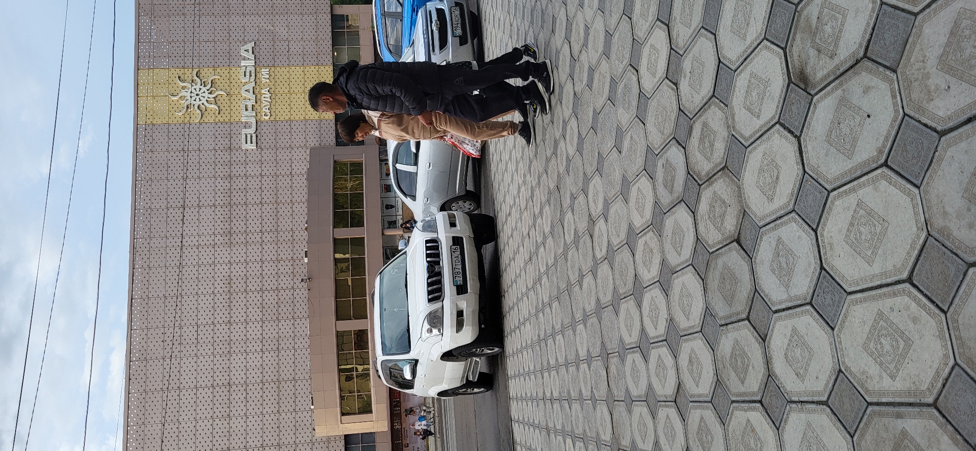 The image captures a street scene in Oskemen, Kazakhstan, in front of the Eurasia shopping center. A white Toyota Land Cruiser Prado is parked on the street, facing the camera. License plate reads "KZ 787 TDA 16". To the right of the Prado, a silver sedan is parked, with its license plate reading "KZ 194 ADR 16". Further right, a blue sedan is partially visible.
Three people are walking from right to left, in front of the cars. The person on the far right is a man in a dark puffer jacket and black pants. Next to him is a woman in a tan tracksuit, carrying a patterned shopping bag. To her left, a man in a black puffer jacket and dark pants walks slightly ahead.
The background features a large, modern building with a beige, tiled facade. The "EURASIA" logo with a sun-like emblem is visible on the upper part of the building. The street is paved with decorative hexagonal tiles in shades of grey and beige. Overhead, power lines crisscross the sky, which is partly cloudy. The scene appears to be a public space, possibly a sidewalk or pedestrian area adjacent to a road.
