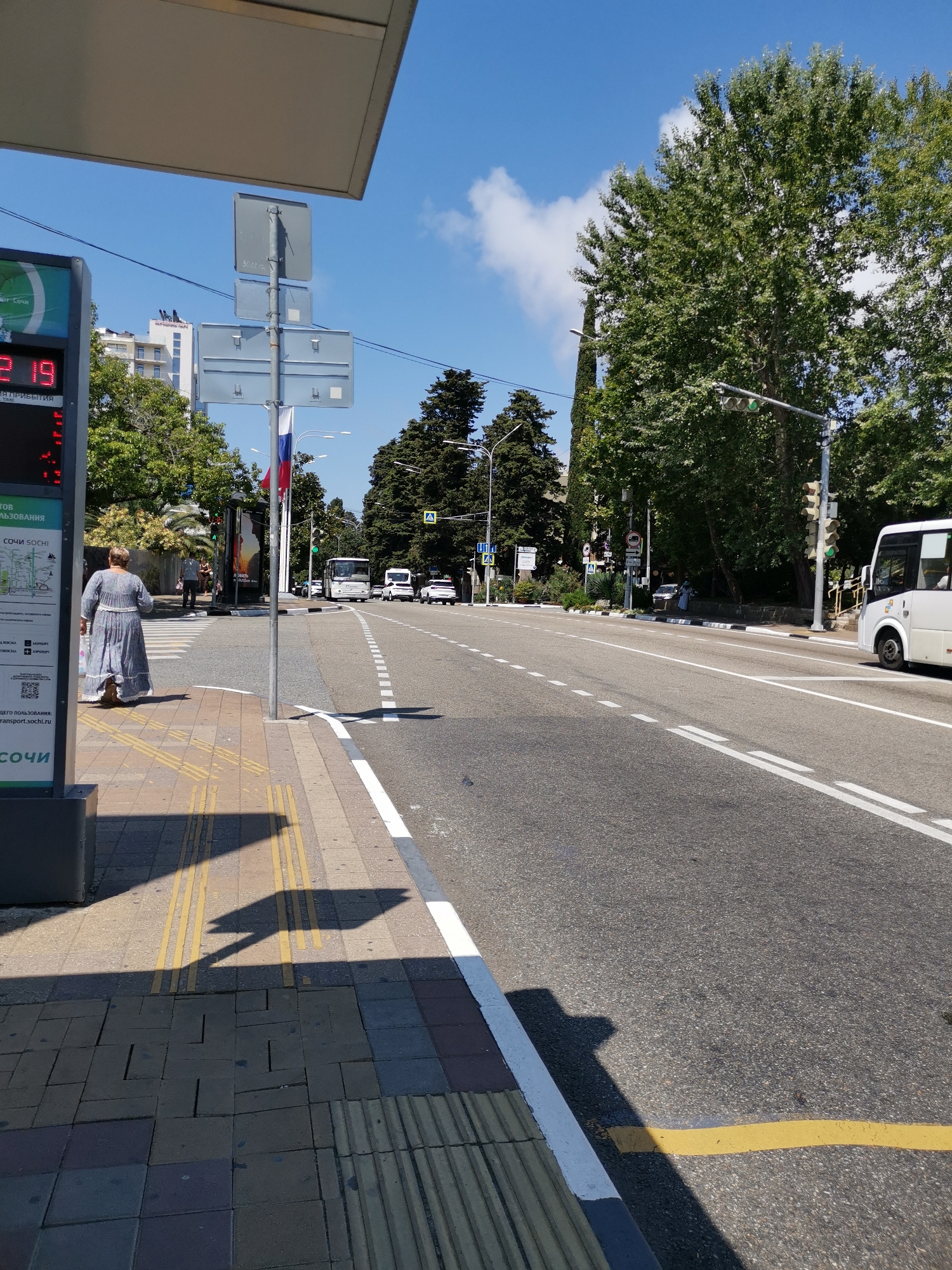 The image depicts a sunny day at a bus stop in Sochi, Russia. On the left side of the frame, a bus stop information kiosk displays route information and has a digital display showing "2.19". A woman in a long, light blue dress is walking away from the kiosk towards the road. Behind her, a few other pedestrians are visible. The road is a multi-lane asphalt surface with clear lane markings. Several vehicles are on the road, including a white bus on the right side and other cars further down. Traffic lights are visible, some showing green. Lush green trees line the sides of the road, and in the background, multi-story buildings and a cypress tree are visible against a bright blue sky with scattered clouds. A Russian flag is attached to a pole near the bus stop. The ground in front of the bus stop is paved with yellow tactile paving for visually impaired pedestrians, and a shadow of a person holding an umbrella is cast across it.