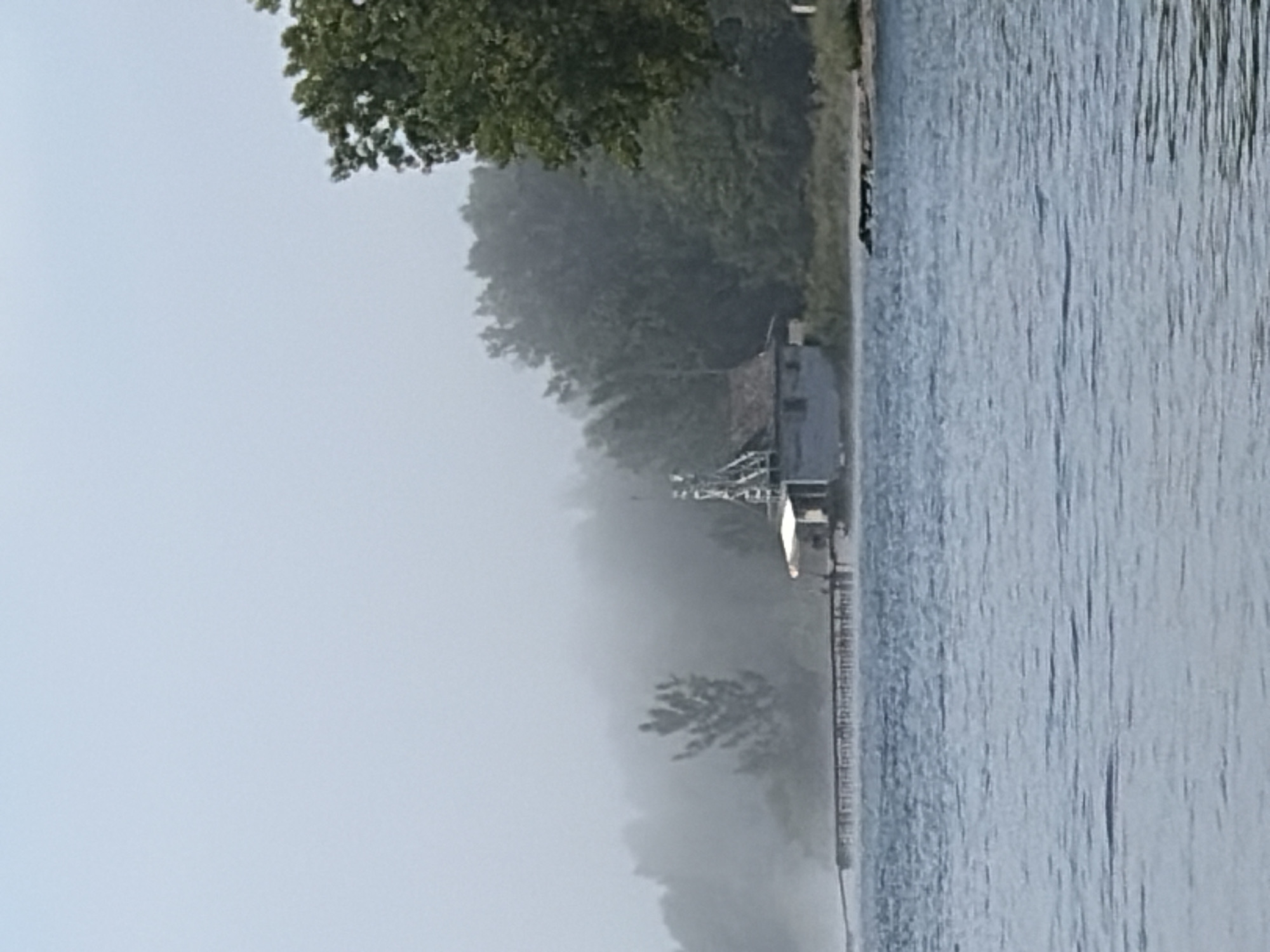 The image depicts a body of water in the foreground, with a shoreline and a building structure visible in the mid-ground. A long pier extends from the left side of the frame towards the building. The building appears to be a small, rustic structure, possibly a boathouse or shed, with a white overhang and a darker roof. It is situated on the edge of a wooded area. The trees are lush and green, suggesting a temperate climate. In the background, a hazy atmosphere obscures the distant landscape, with the sky appearing light and overcast. There are no people or vehicles visible in the scene. The overall impression is one of a quiet, natural setting, possibly a rural or park-like area.