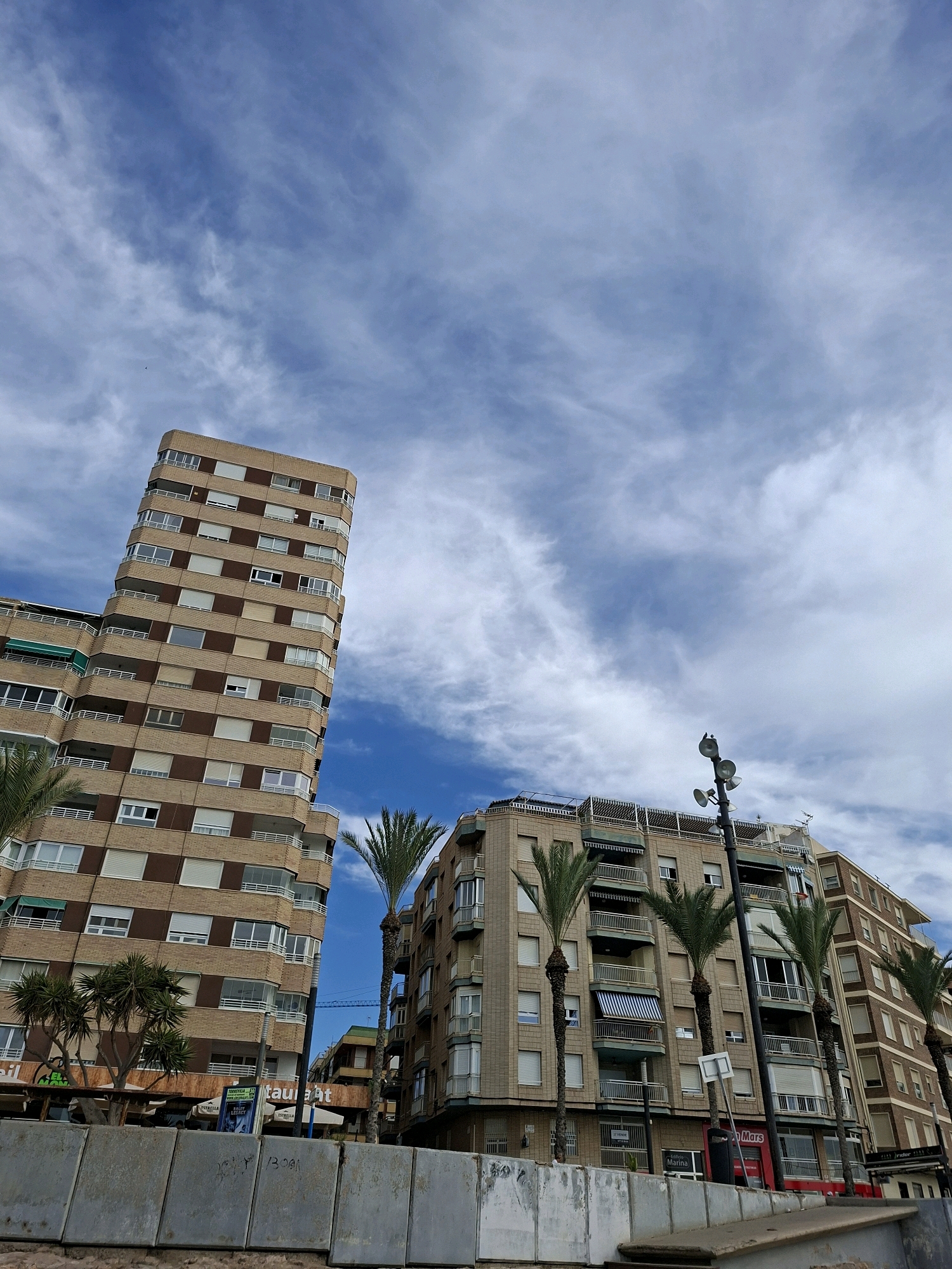 An exterior, low-angle shot captures two multi-story apartment buildings and a street lamp against a partly cloudy blue sky. The building on the left is taller and features a rounded facade with rows of balconies and windows. The building on the right is shorter and also has balconies and windows. Palm trees are visible in front of both buildings. At the base of the buildings, storefronts are visible, including one with "Restaurant" in orange lettering. In the foreground, a concrete barrier with graffiti runs along the bottom of the image. The scene is set in Torrevieja, Spain, a coastal city known for its beaches and vibrant atmosphere.