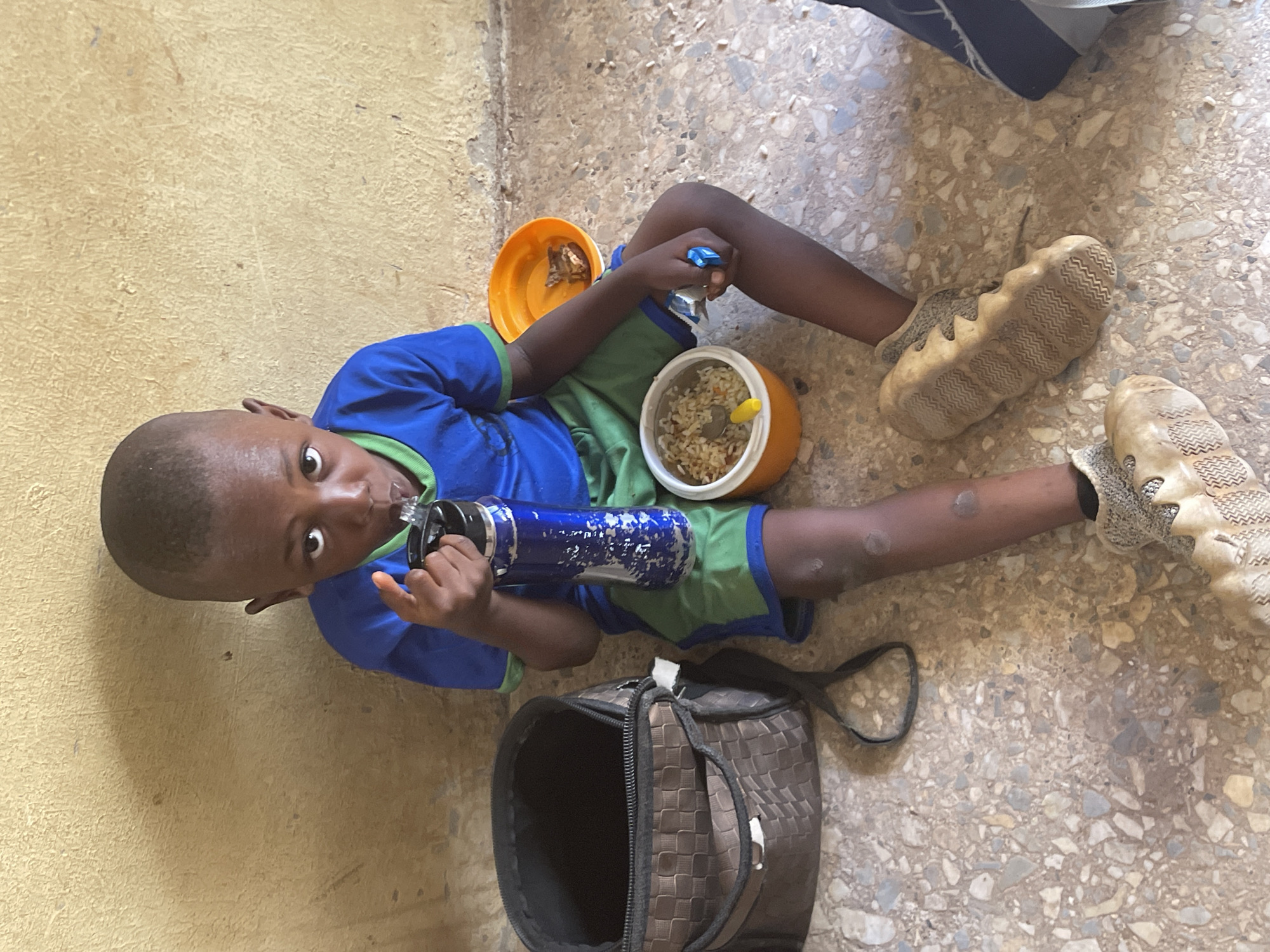 The image, taken in Akwanga, Nigeria, captures a young child lying or sitting on a light-colored, mottled terrazzo or concrete floor, looking directly at the camera. The child, with dark skin and short hair, is dressed in a blue short-sleeved shirt with green trim and green shorts. He is wearing light brown or beige sneakers with textured soles.
In the center of the frame, the child is holding a blue and silver patterned water bottle to his mouth, appearing to drink. Two orange plastic bowls are placed on the floor near him. One bowl contains a meal of rice mixed with vegetables, along with a small yellow plastic spoon. The other orange bowl holds a smaller quantity of food, possibly meat or another dish.
A black and brown checkered soft-sided bag, open at the top, is visible on the floor near the child's legs. A small blue object, possibly a pen or a toy, lies near his right hand. The background consists of a plain, textured light brown or beige wall. The lighting suggests it is daytime, likely indoors or in a shaded area. The child's expression is calm and inquisitive, with wide eyes. No discernible text is visible in the image.