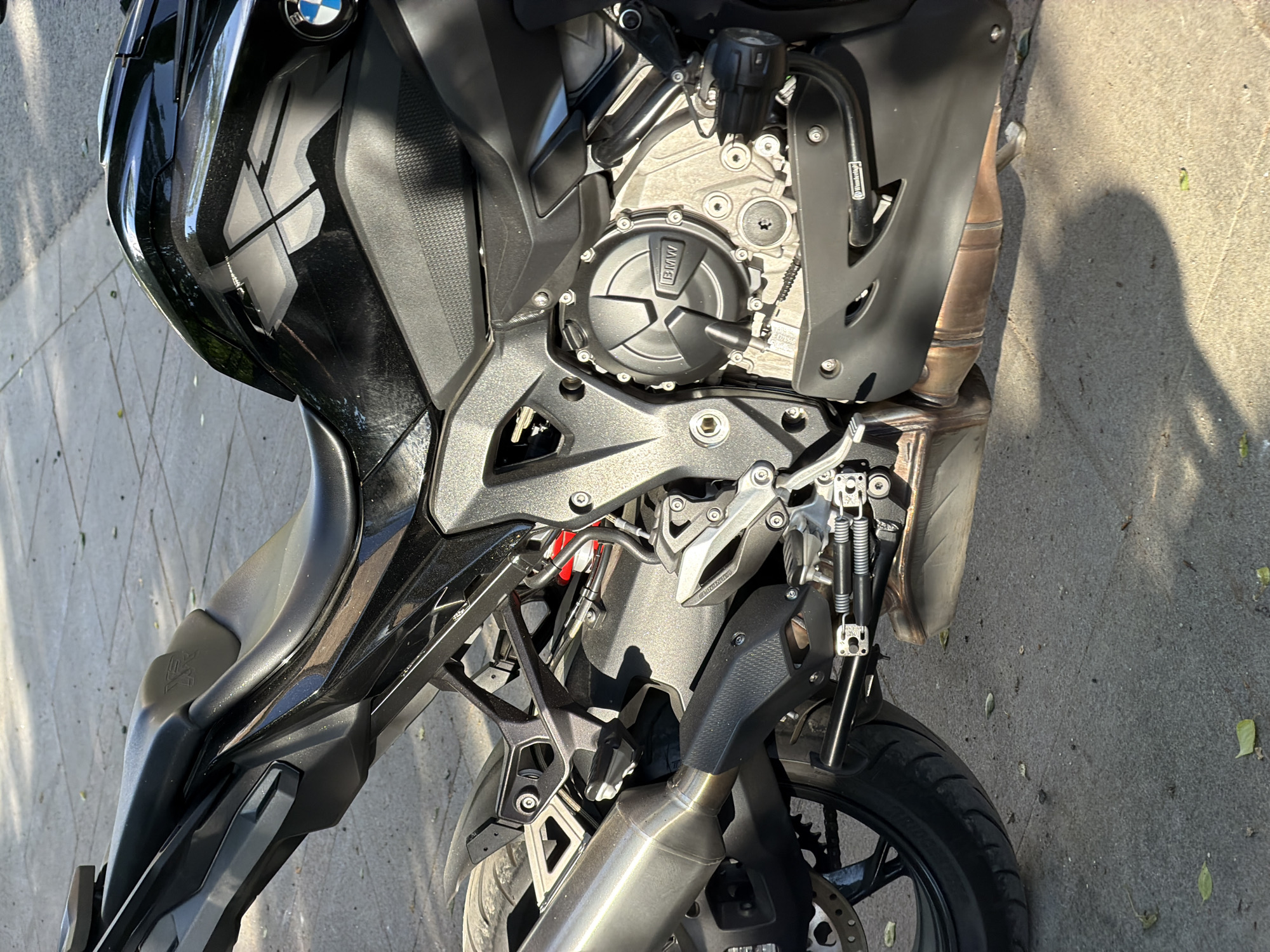 The image shows a close-up of the left side of a black BMW motorcycle parked on a concrete surface, likely a sidewalk. The motorcycle's engine, frame, exhaust, and front tire are visible. The BMW logo is prominently displayed on the engine casing. The general location appears to be outdoors on a sunny day, based on the shadows. There's no indication in the image that the location is Moscow, Russia. No people are visible in the frame.