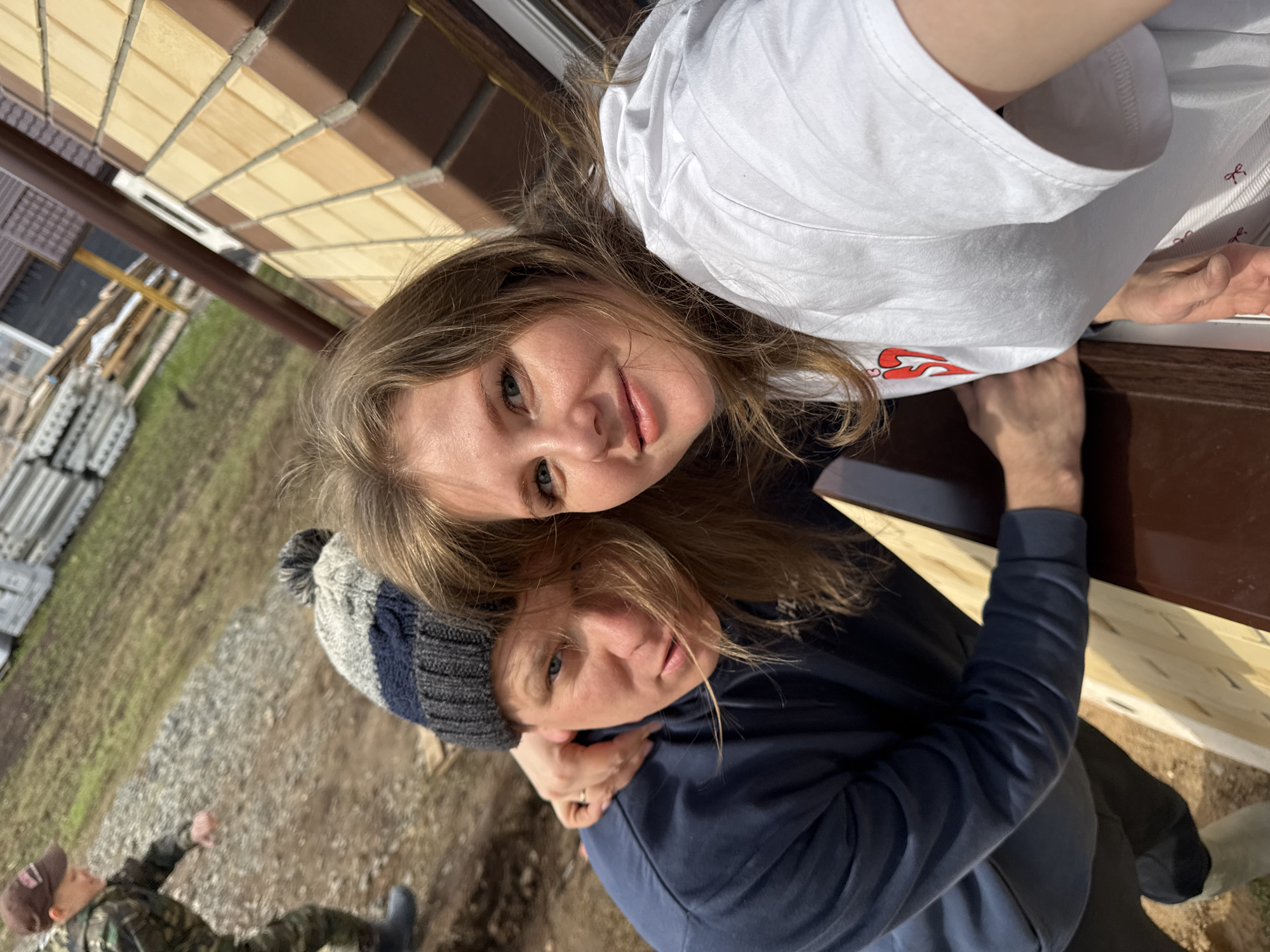 The image shows two individuals, likely women, posing closely together. The woman in the foreground wears a navy blue sweatshirt and a gray and blue knit hat with a pompom. The woman in the background wears a white T-shirt and has long, light brown hair. They are standing near a building with tan and brown siding. In the background, there is a glimpse of a yard with gravel and what appears to be a building under construction. A figure is visible in the lower left corner, wearing military camouflage. The location is likely in a rural or suburban area, based on the construction and terrain. The city mentioned, Gornyi in Russia, cannot be confirmed based on the visible content, as there are no identifiable landmarks or signs present.