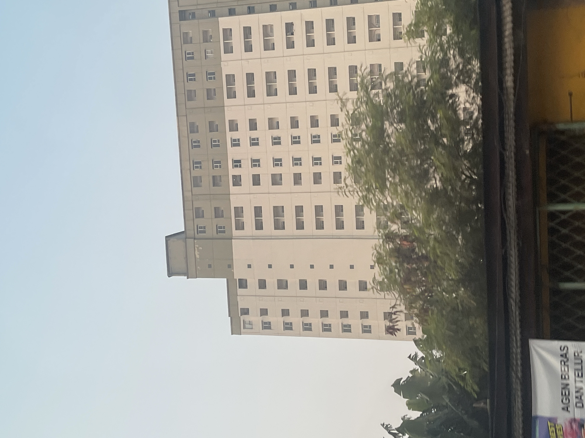 This is a vertical, eye-level, medium-shot photograph of a tall, multi-story apartment building, likely located in the city of South Tangerang, Indonesia. The building has a light-colored facade with rows of dark-framed windows. The upper portion of the building is a different color, appearing to be a pale green or gray. Trees with lush green foliage obscure the lower part of the building. A dark, corrugated roofline is visible at the very bottom of the frame, with a banner hanging from it. The sky above is a pale, hazy blue. No people are visible in the image. The scene appears to be a residential or mixed-use urban environment.