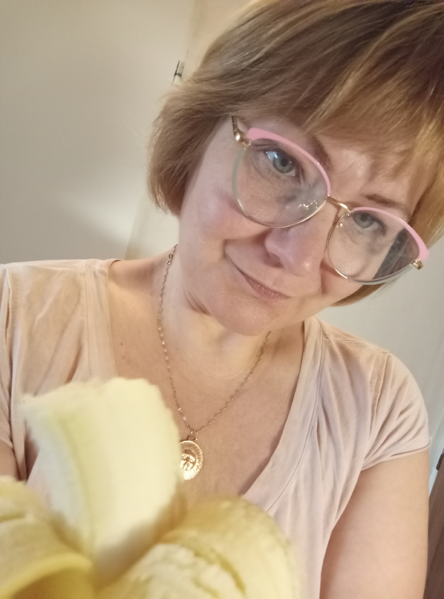 A woman with short blonde hair and blue eyes is looking down at the camera. She is wearing glasses with pink and gold frames and a light pink t-shirt. A gold necklace with a circular pendant is visible around her neck. In the foreground, a partially peeled banana is being held up, obscuring a portion of her body. The background appears to be an indoor setting with neutral-colored walls. The image is a close-up, self-taken photograph.