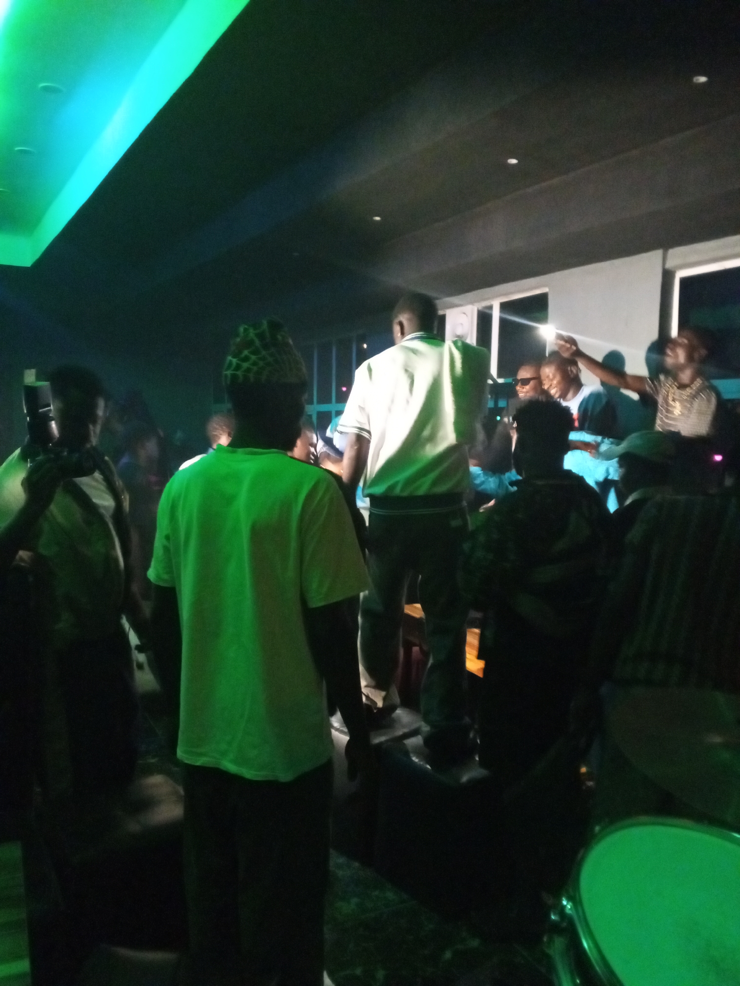 A nighttime indoor scene. A group of people gather, with one person standing on a raised platform or speaker. A man stands in the foreground wearing a green shirt and a patterned cap. Another person holds a camera. There is a drum kit in the foreground. The lighting is dim and green, with green light fixtures on the ceiling. The location is likely a bar, club, or performance venue. The city, Lokoja, Nigeria, is based on your given information.