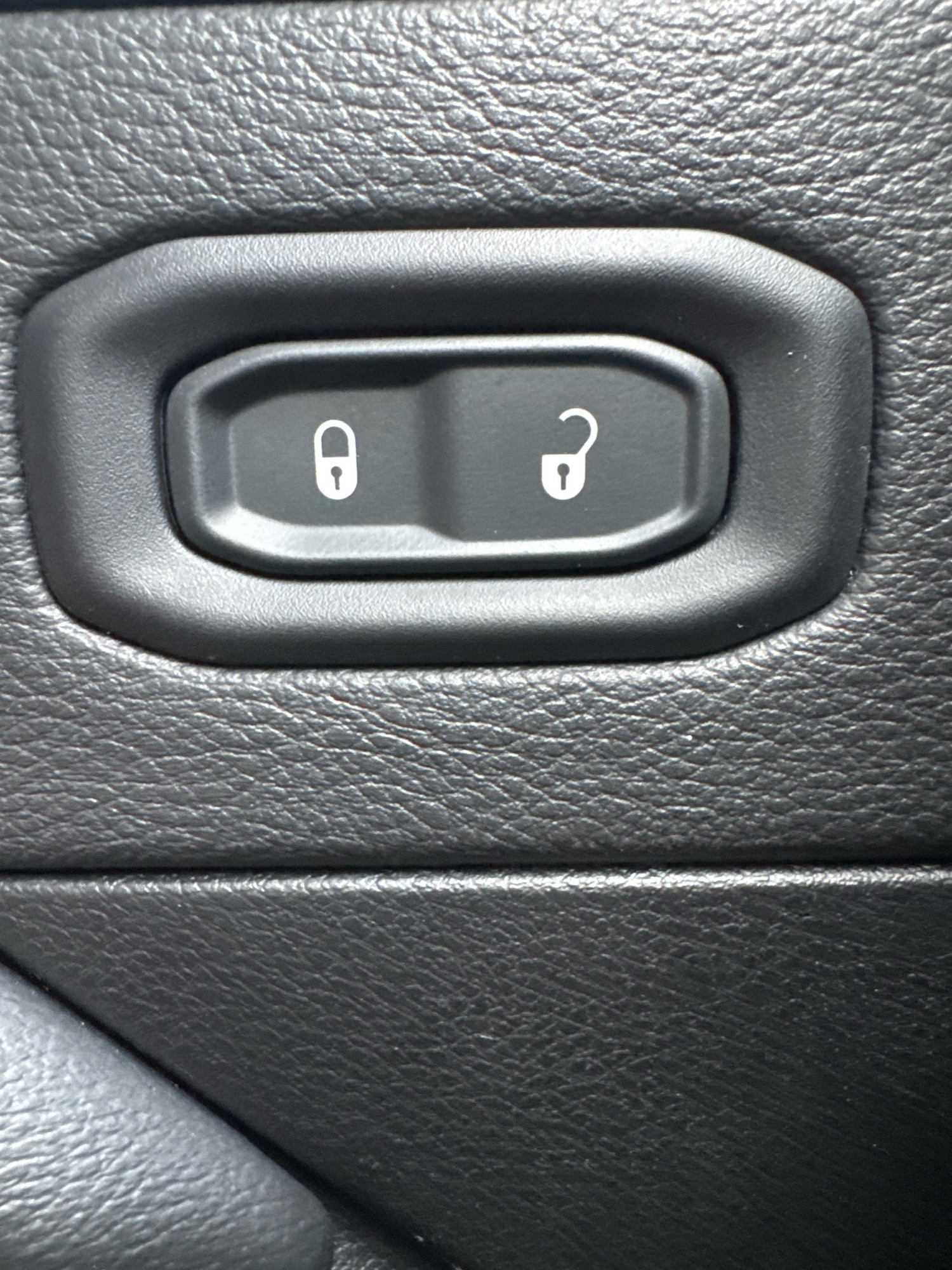 A close-up shot of two car door lock buttons on a textured black surface. The button on the left displays a white icon of a locked padlock, and the button on the right shows a white icon of an unlocked padlock. The buttons are inset into a rounded black plastic housing. The background is a black, textured material, likely leather or a synthetic imitation, with a visible grain pattern. The lighting suggests an interior car environment.
