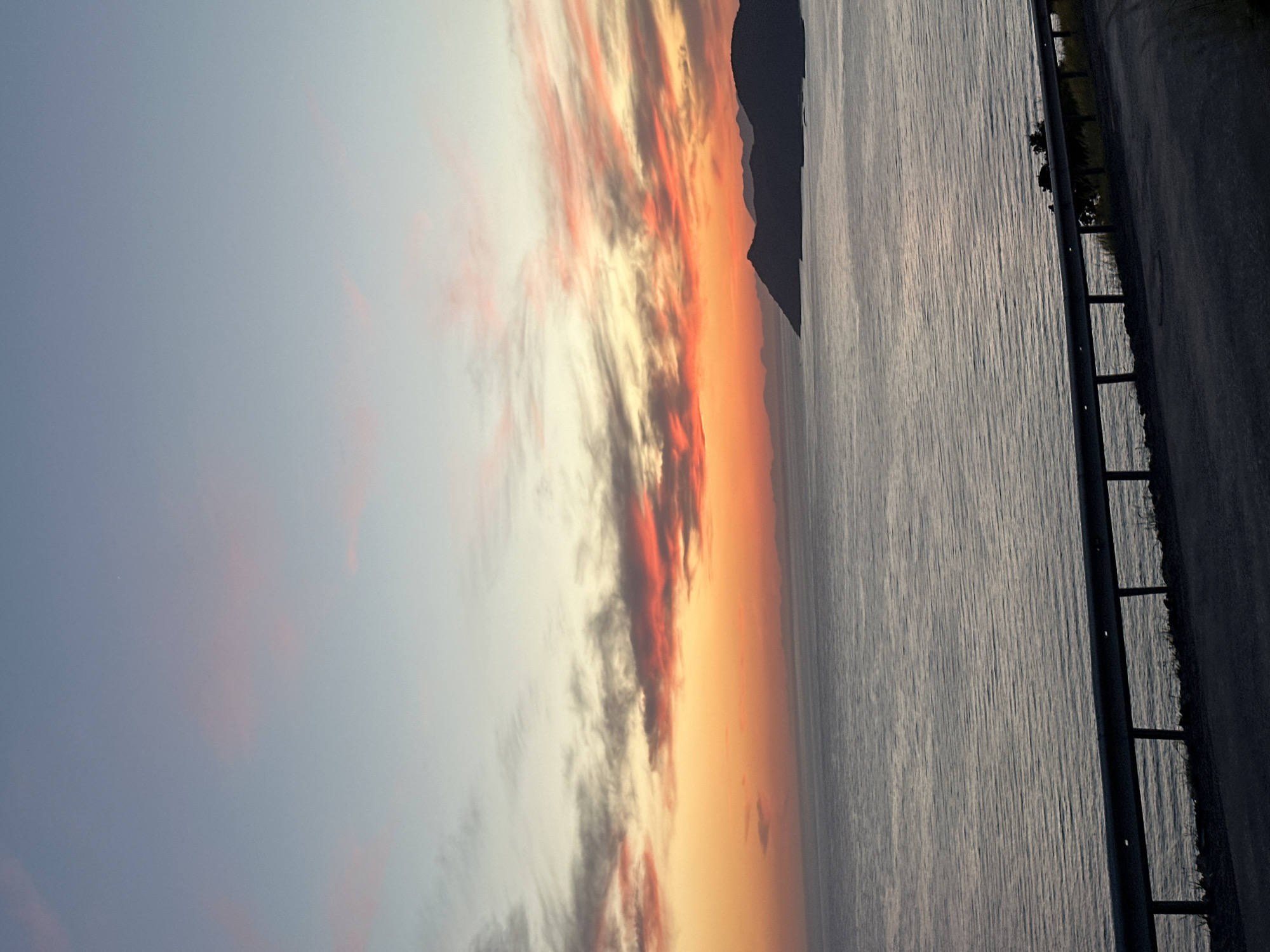 The image, correctly oriented horizontally, captures a vibrant coastal landscape at either sunrise or sunset. The sky is a dominant feature, filled with dramatic, elongated clouds displaying a striking palette of colors, ranging from fiery orange and deep red to soft yellow and cooler grey and light blue. These colorful streaks stretch across the upper part of the frame, creating a captivating celestial display.
On the distant horizon, a dark, undulating silhouette of mountains or hills separates the vivid sky from the expansive body of water below. The water, likely the sea, appears calm with subtle textures and reflections, taking on a metallic, silvery-grey hue.
In the right foreground, a paved road or pathway runs parallel to the water. A dark, sturdy metal guardrail with vertical posts separates the road from the coastline, suggesting a scenic drive or viewpoint. Small areas of sparse green vegetation are visible between the guardrail and the water's edge.
The warm, intense hues in the sky strongly indicate the time of day is either dawn or dusk, with sunset being a probable interpretation given the depth of colors. The scene evokes a sense of tranquility and natural beauty, consistent with coastal regions like Fethiye, Turkey. There is no visible text in the image.