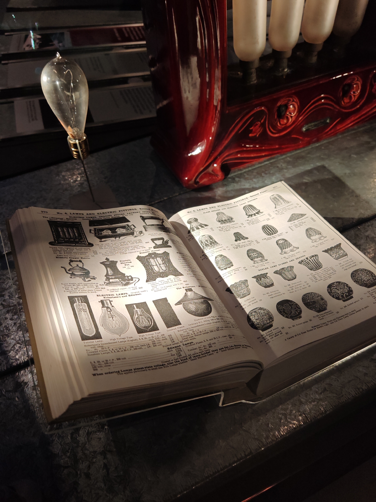 A close-up shot shows an open book titled "No. 4. LAMPS AND ELECTRIC FITTINGS" on a textured surface. The book displays illustrations and descriptions of vintage electric lamps, shades, and related items. Adjacent to the book is a vintage-style light bulb. A red ornamental structure with what appears to be milk glass tubes sits in the background. The setting appears to be a museum or exhibit space showcasing antique lighting technology. There is no indication that this is in the city of London, United Kingdom.