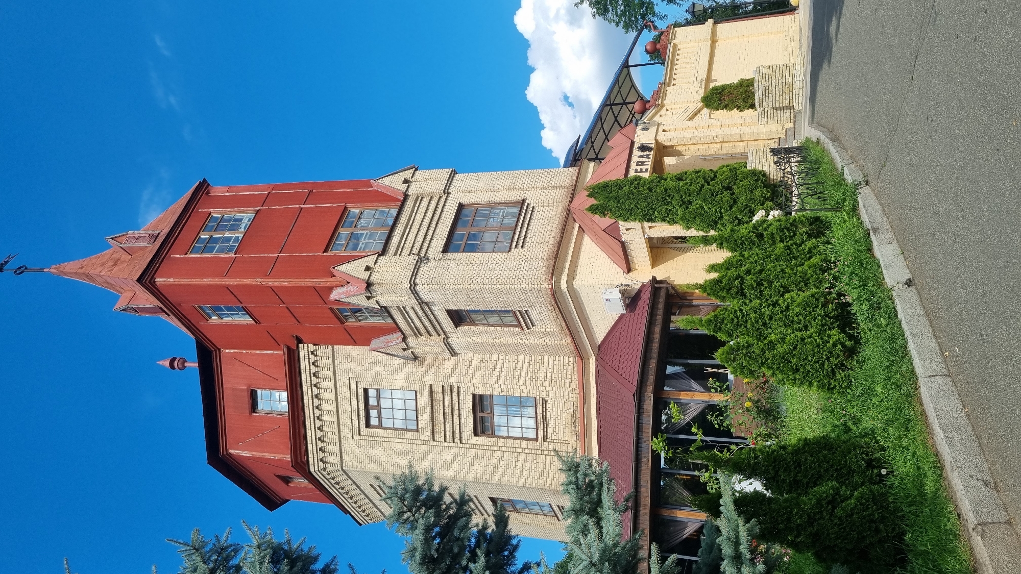 The image depicts a building with a distinctive red, pointed roof and a tall, red, multi-story structure attached to a lighter-colored brick building. The building is situated outdoors, with a clear blue sky and some white clouds visible. Green trees and bushes surround the building, with one section featuring manicured hedges and a paved walkway. A road runs alongside the grassy area, bordered by a curb. The overall scene suggests a daytime setting under fair weather.
The building's architectural style is a blend of traditional and possibly historical elements, with the red tower resembling a watchtower or a unique design feature. The yellow brick section of the building has multiple windows and architectural details, including decorative brickwork. The foreground shows lush greenery, suggesting a well-maintained outdoor space, possibly a garden or park area adjacent to the building.
There is visible text on the building, above an entrance, which reads "ERA". The context of Kyiv, Ukraine, is provided, which might indicate the building's location or purpose within the city. No people are visible in the image, and no specific activity or event is clearly taking place, other than the general depiction of a building and its surroundings. The lighting suggests it is daytime, likely a sunny day given the bright sky.