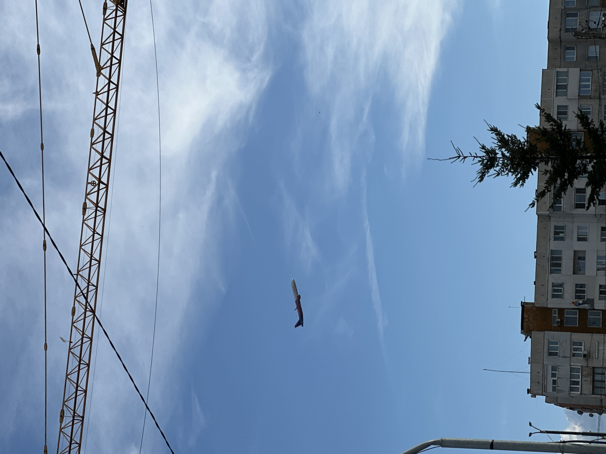 The image, taken in Sofia, Bulgaria, shows a blue sky with cirrus clouds. A large construction crane is visible on the left side of the frame, its orange structure contrasting with the sky. In the center, a plane with red and blue markings is flying vertically. On the right, a tall, concrete residential building with numerous windows is partially obscured by a tree. A street light fixture is in the lower right corner.