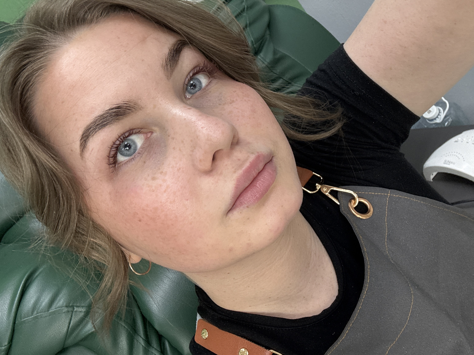 A close-up shot depicts a fair-skinned woman with light brown, wavy hair and blue eyes. She has freckles on her face. She is wearing a black top under a gray apron with brown straps and gold hardware. A small gold hoop earring is visible. She is lying on a green, possibly leather, cushioned surface. The background includes a white object, potentially a nail lamp. The location is possibly in Chaikovsky, Russia, but this is unconfirmed from the image.