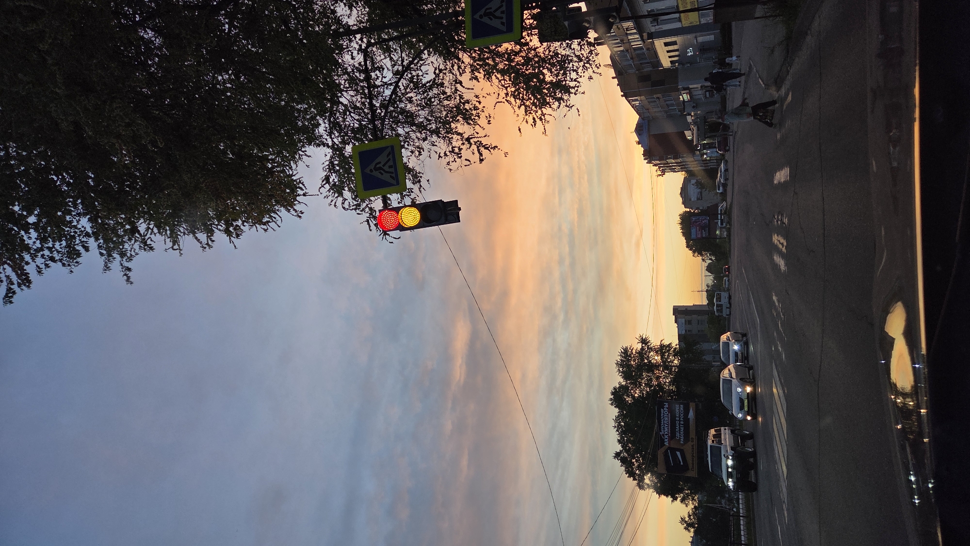 The image depicts a street scene in Birobidzhan, Russia. A traffic light showing red and yellow is suspended above the street near pedestrian crossing signs. The sky is a mix of orange and blue, likely during sunset or sunrise. Buildings line the side of the street. Several cars are seen in the street. Trees are present in the foreground and background. The photo appears to be taken from inside a vehicle.