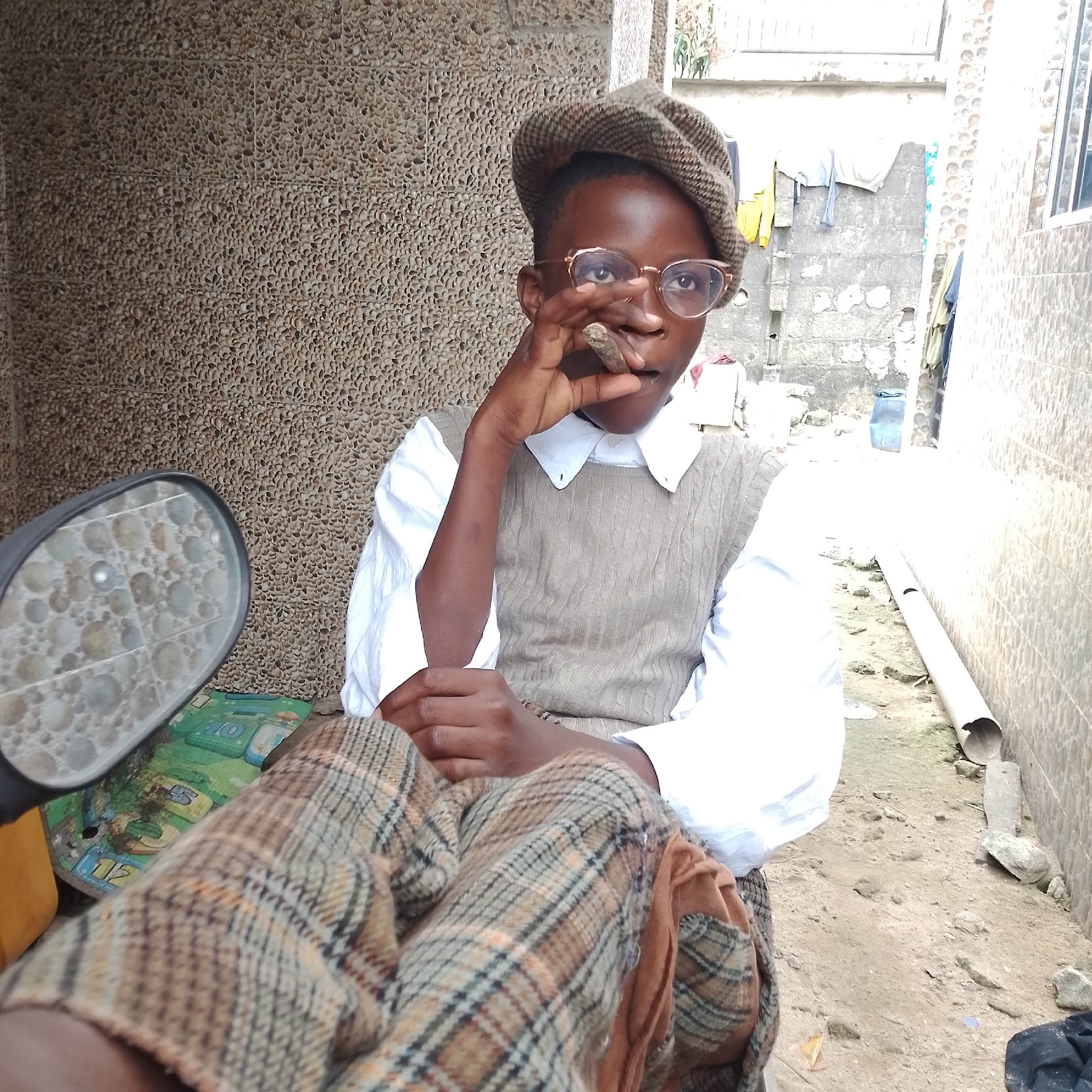 In Ado, Nigeria, a young person is photographed indoors, seated and holding a cigar to their lips. They are wearing a brown and tan plaid cap, gold-rimmed glasses, a white collared shirt, and a tan cable-knit vest. Their legs are crossed, covered by brown and tan plaid pants. The background consists of a textured, pebbled wall, a dirt ground, and some pipes and miscellaneous items. The lighting suggests it is daytime.