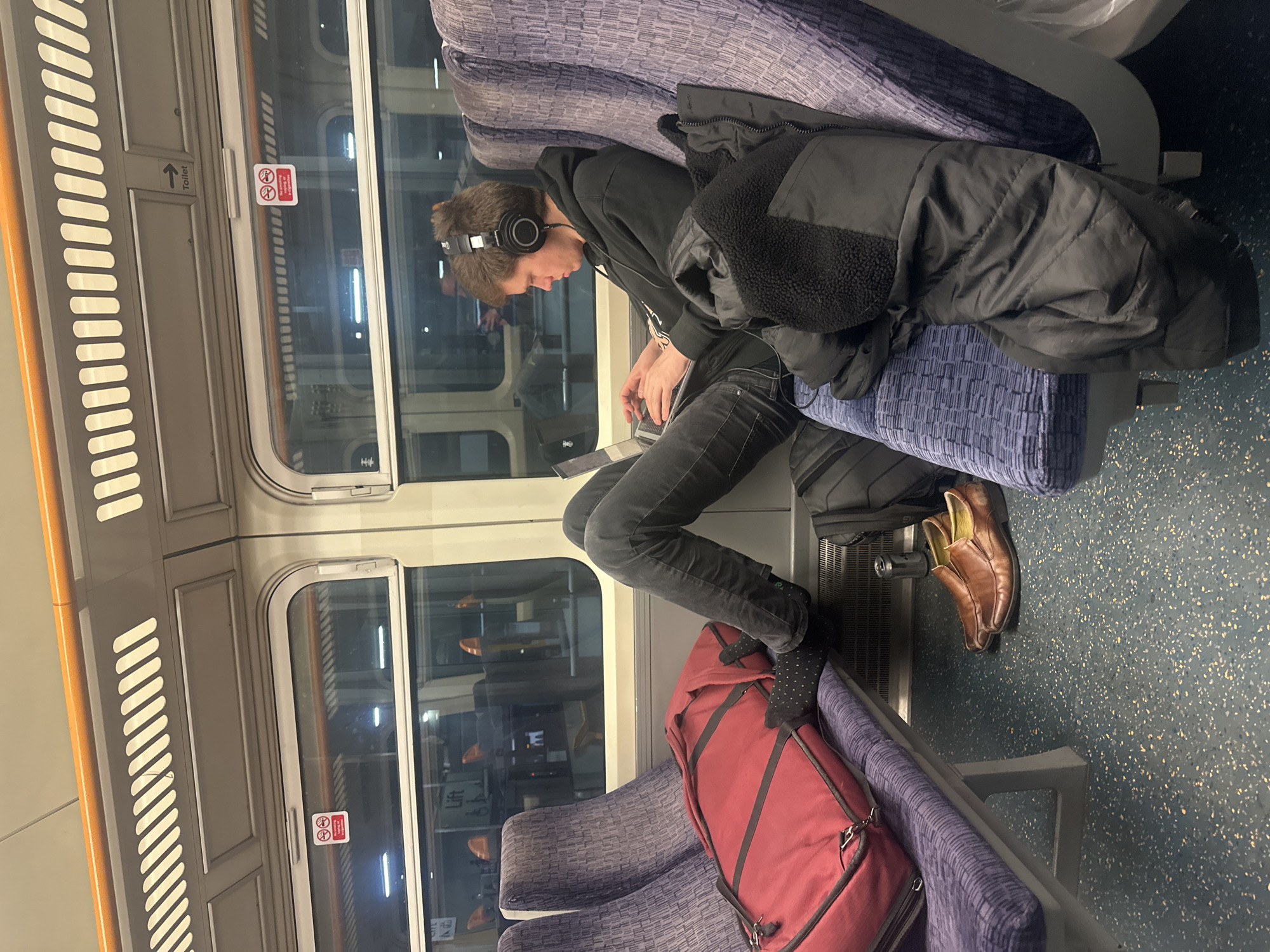 The photograph depicts a person seated on a train. The individual wears headphones and is using a laptop. A black jacket rests on the seat next to them, and a red duffel bag sits on the adjacent seat. The train interior features blue patterned seats and large windows. Based on the interior design of the train it is highly likely to be within the Transport for London network of London, United Kingdom.